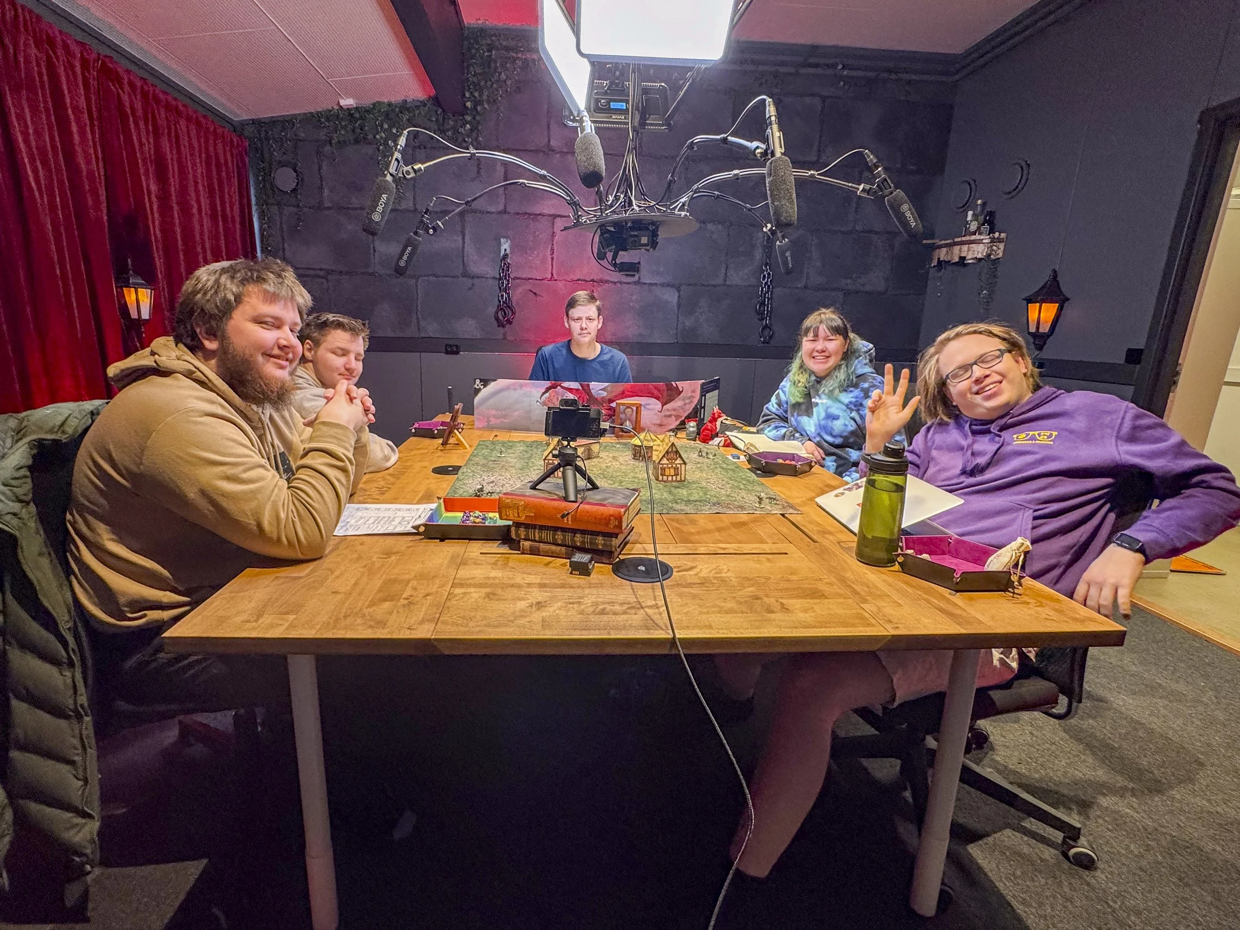 Fem personer rundt et bord med rollespillutstyr, lommekamera på stativ, mikrofoner henger i taket, røde og svarte vegger, en person vinker og smiler, andre ser oppmerksomme ut.