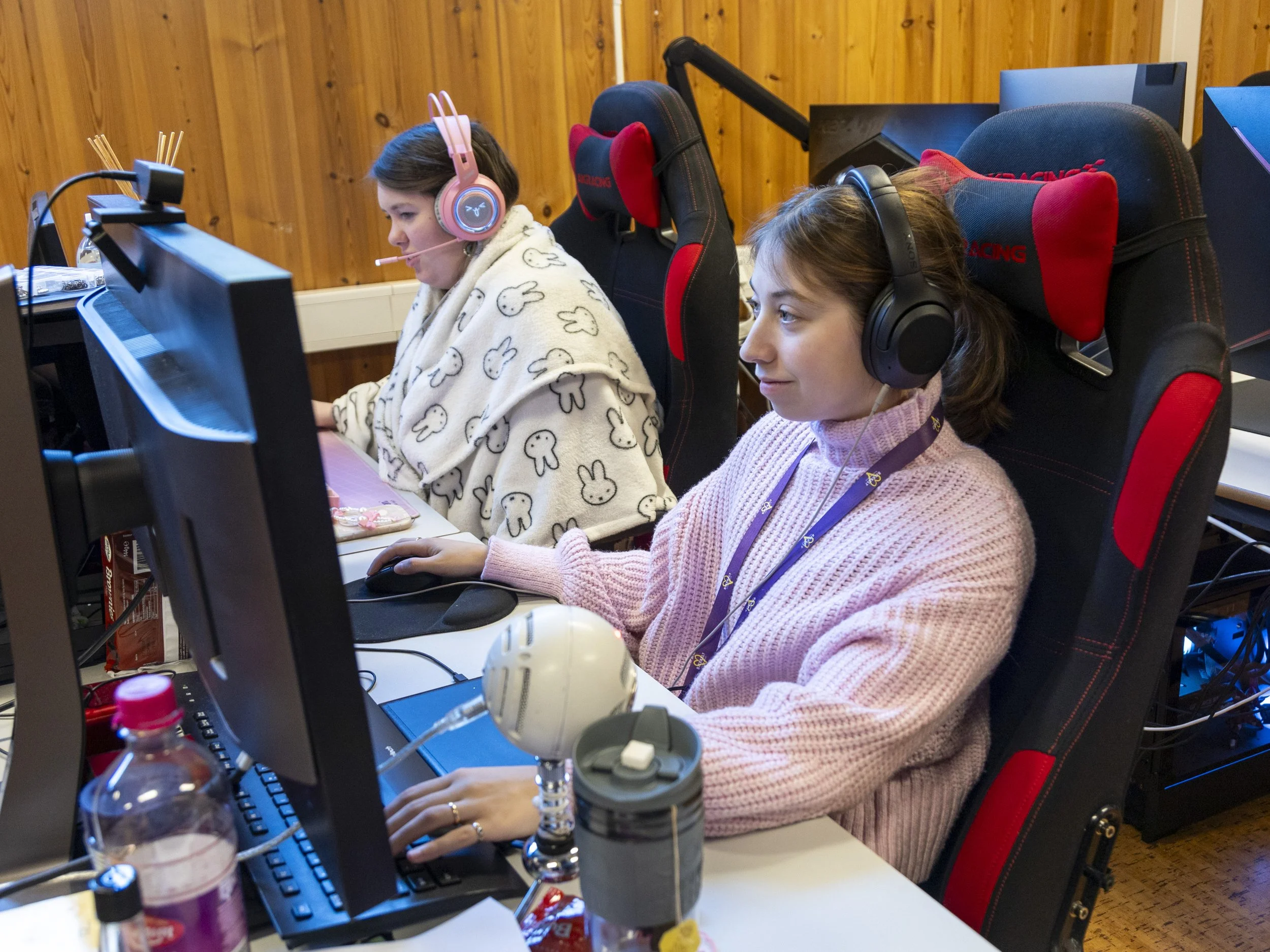 To unge jenter som spiller dataspill i et rom med tre datamaskiner og headset.