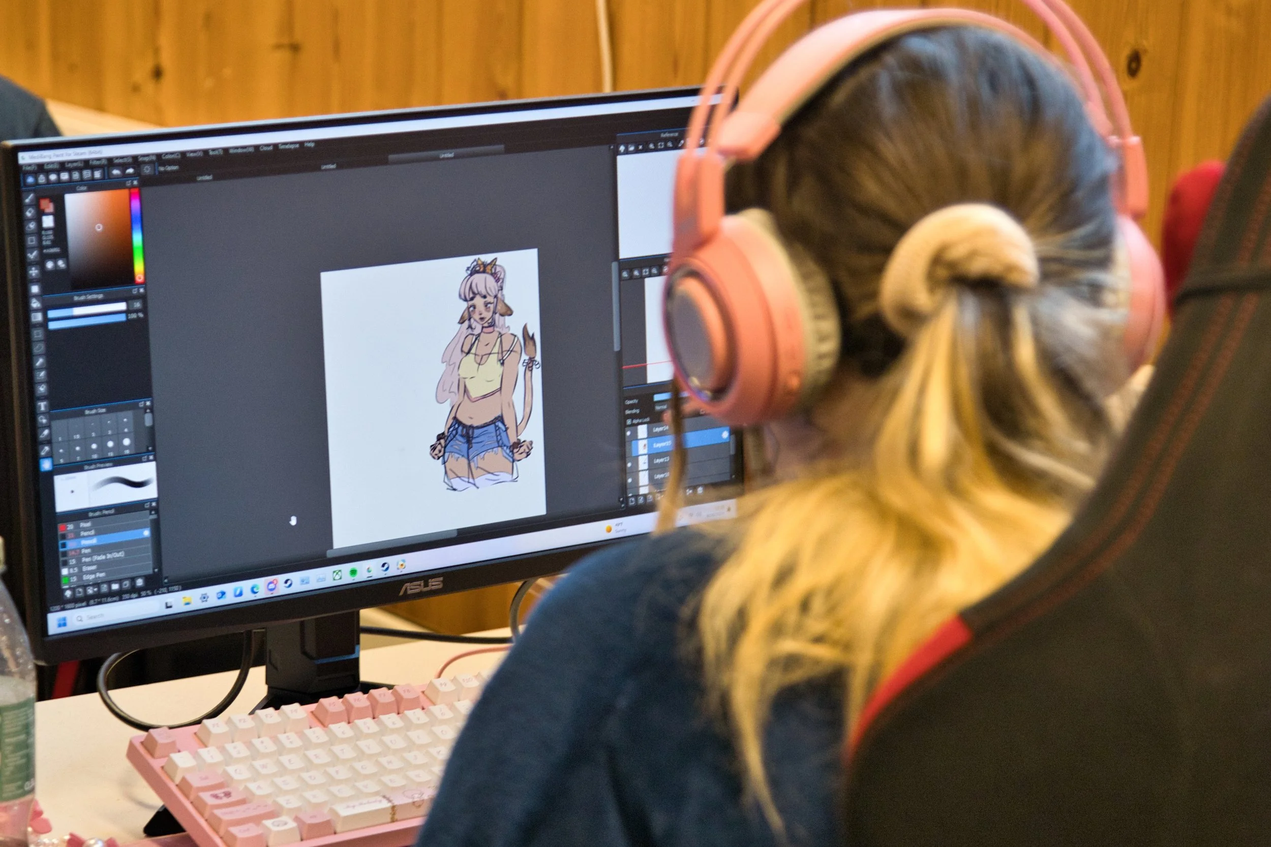 En person med langt blonde hår og rosa hodetelefoner som jobber med en illustrasjon på en dataskjerm, hvor det vises en tegning av en karakter med katteører og hale.