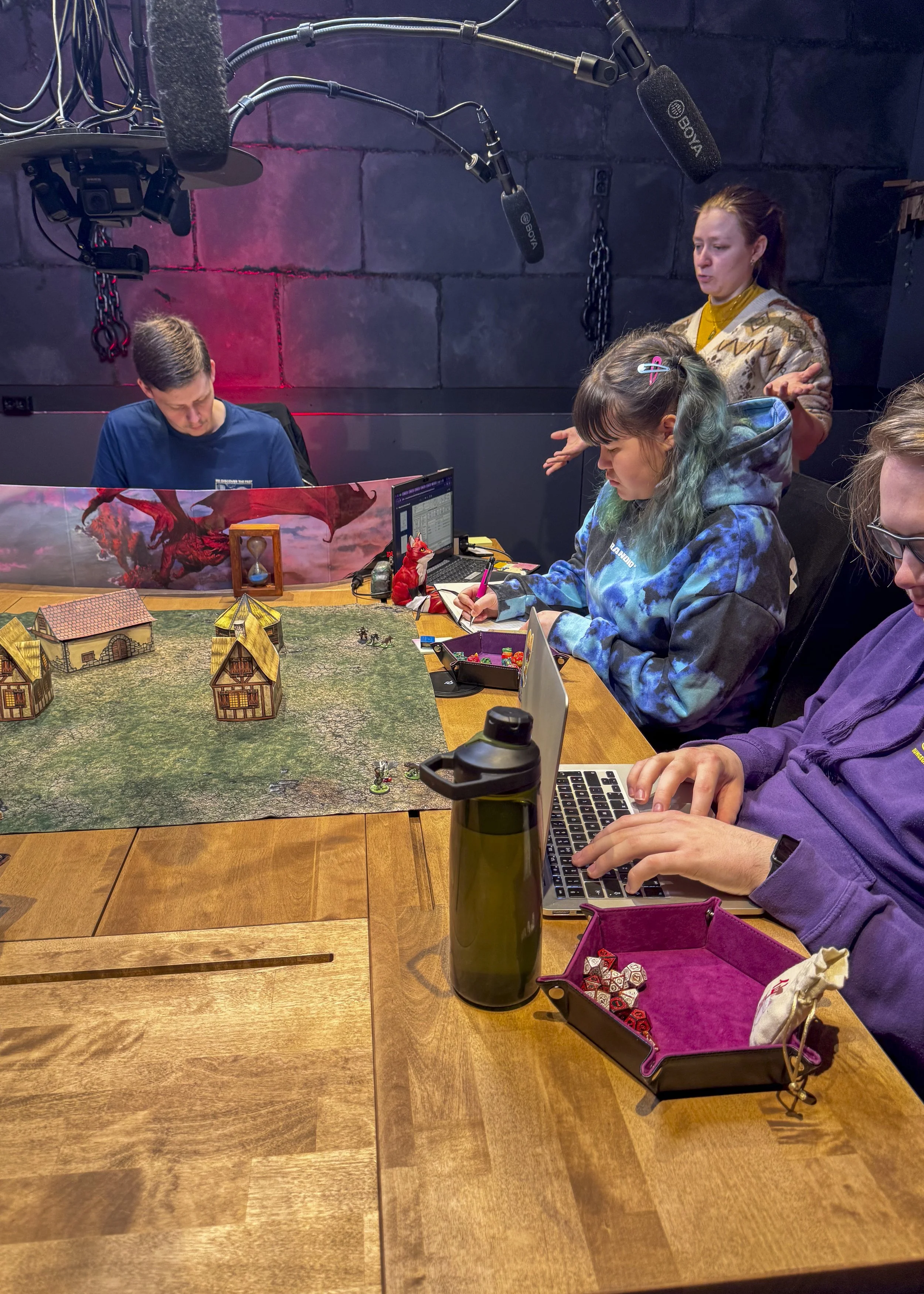 Gruppe med fire personer som spiller bordrollespill med en miniatureverden på bordet, omgitt av kamerautstyr for innspillingsformål.