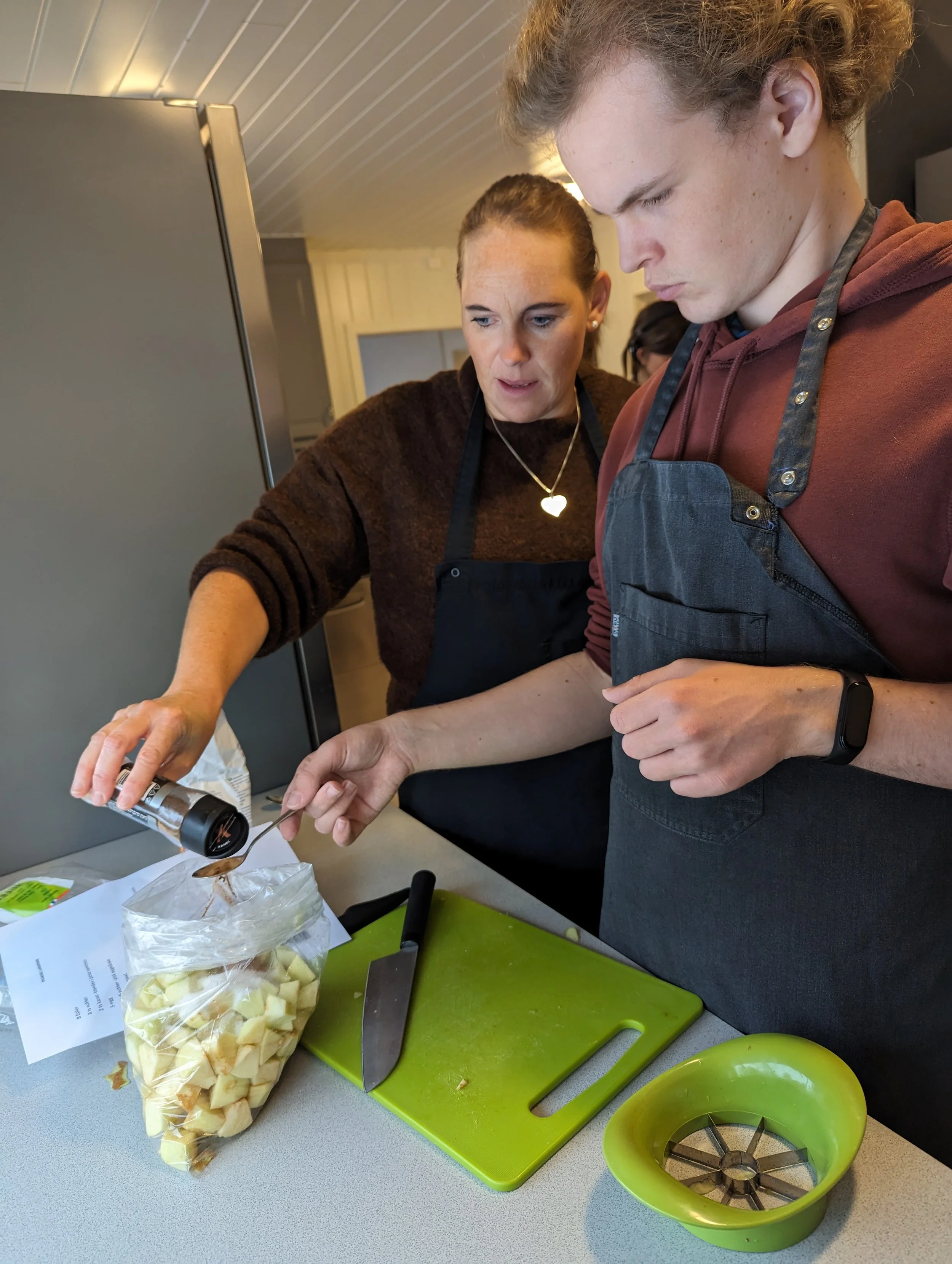 To personer som lager mat i et kjøkken. Den ene person fyller en plastpose med oppkuttet eple, mens den andre bruker en krydderspiral og har et grønt skjærefjøl og en grøn skive.