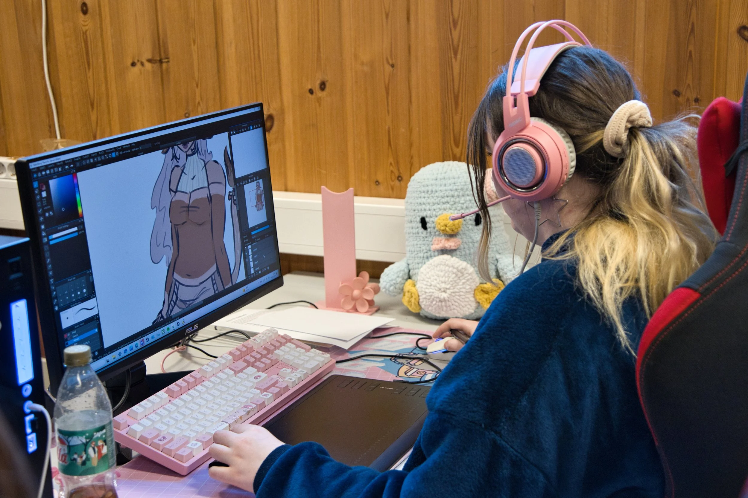 Person med rosa hodetelefoner som jobber på datamaskin med illustrasjoner av en kvinnelig karakter på skjermen, omgitt av stuffed dyr og dekorasjoner.