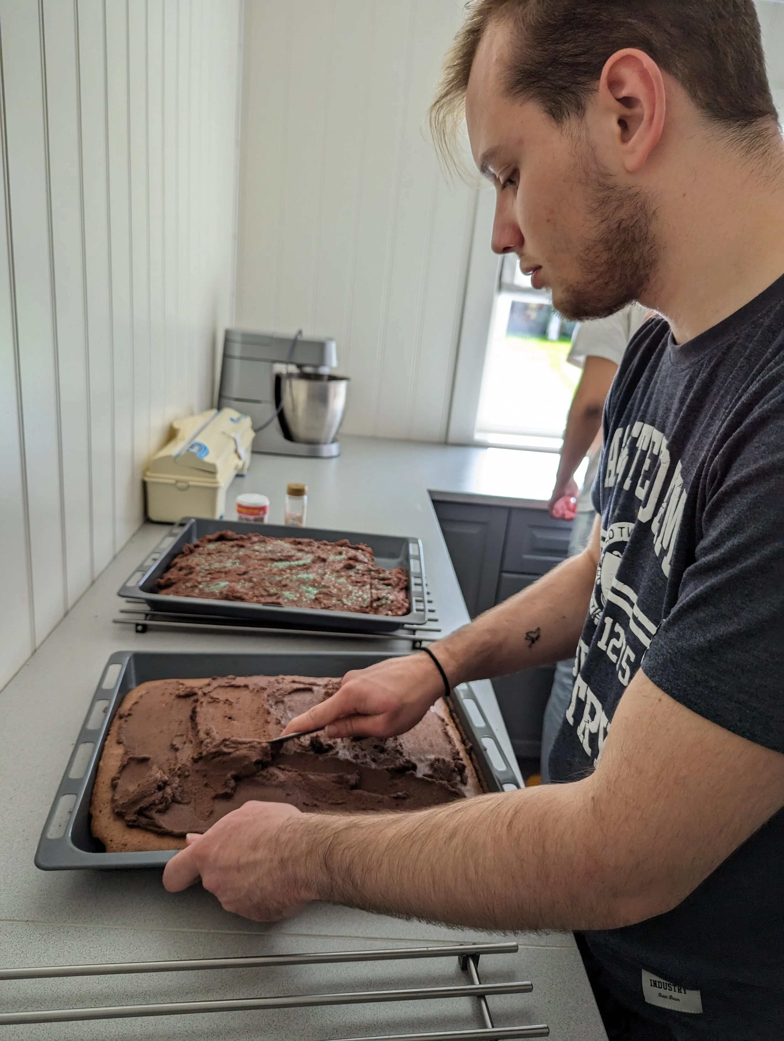 En person med blond hår og skjegg redegjør i en bakker med sjokoladekake, mens andre personer står i bakgrunnen.