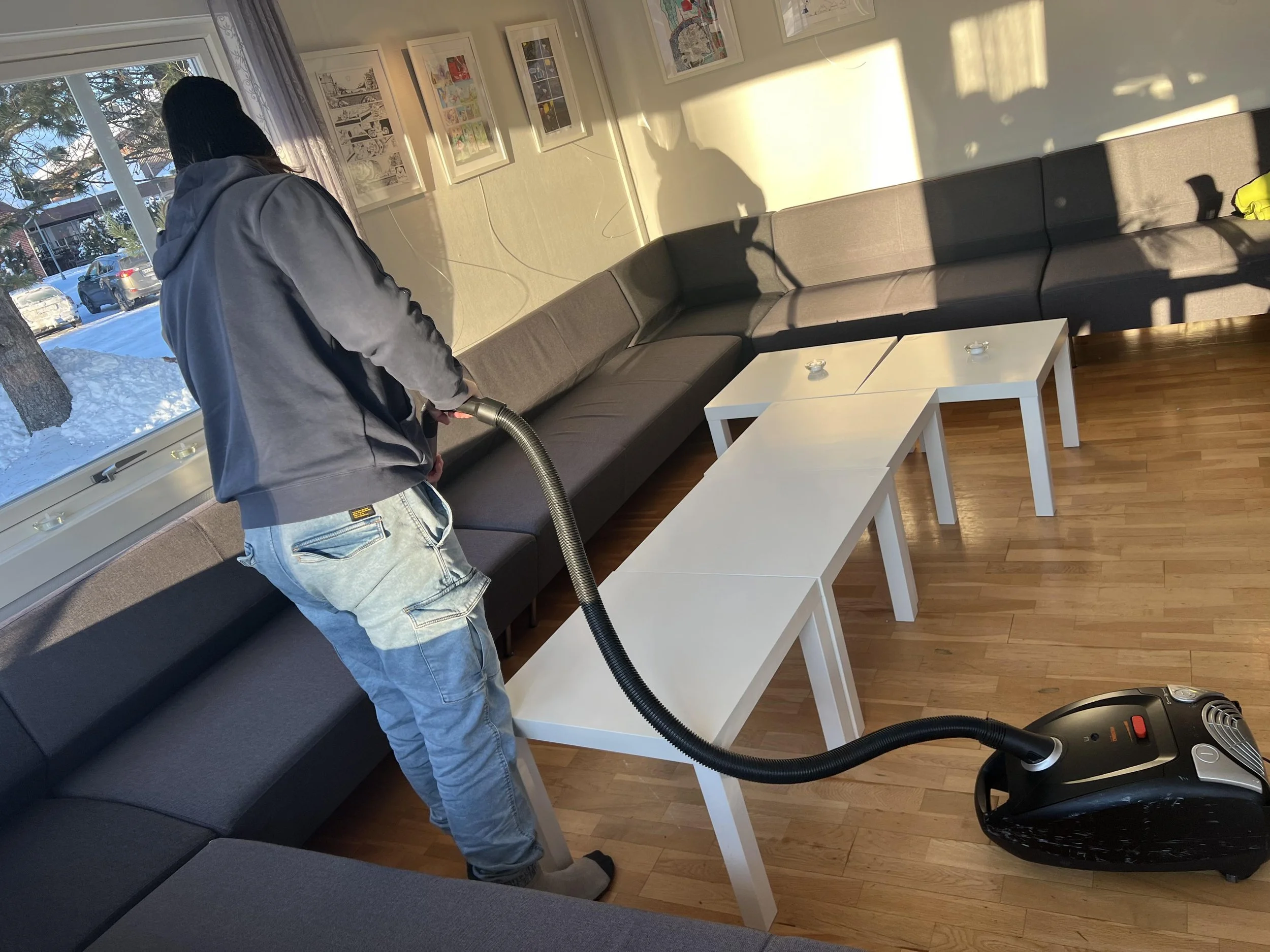 En person bruker støvsuger på en sofa i et rom med flere kvadratiske hvite bord og veggdekor.