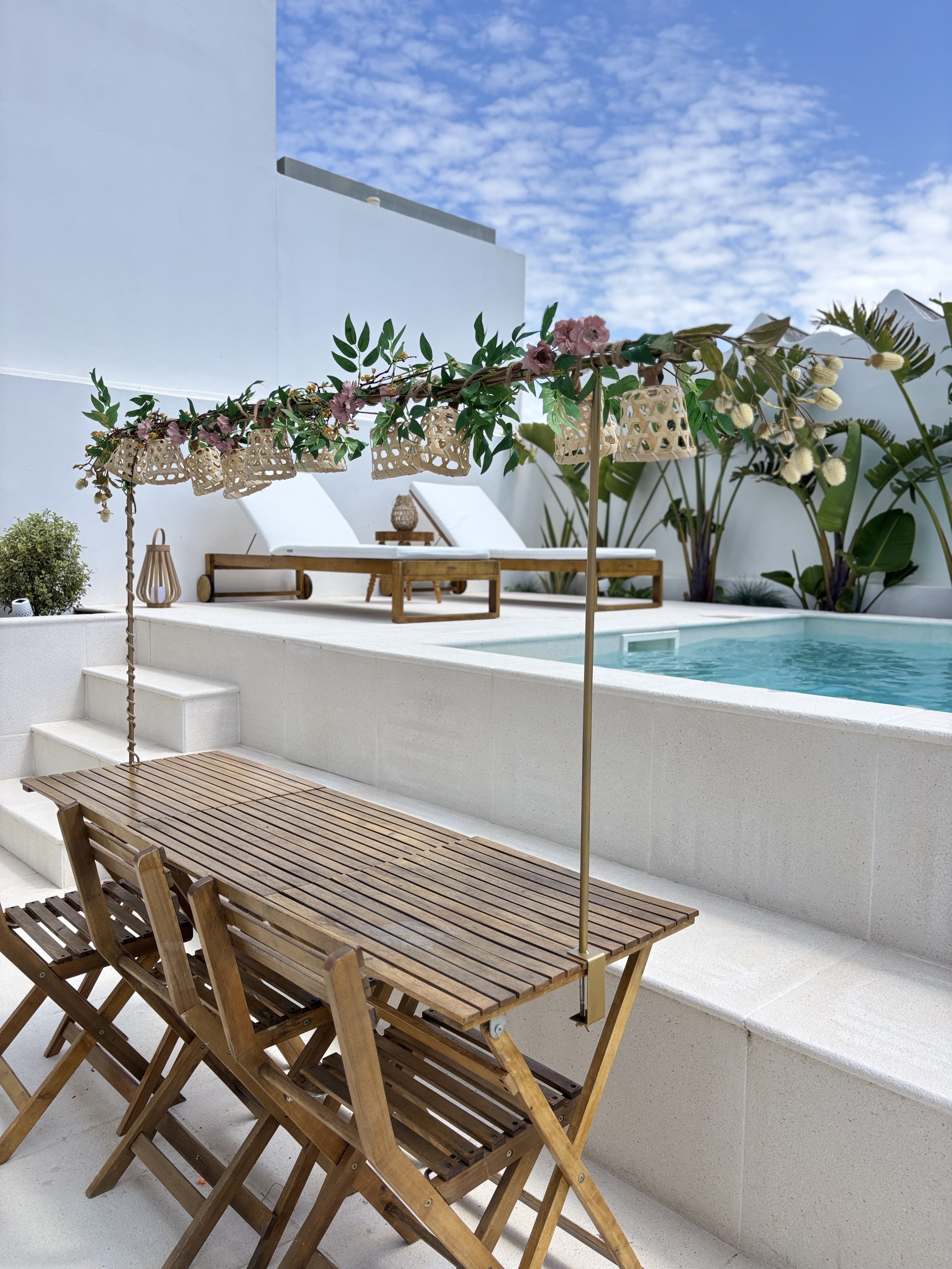 Outdoor pool area with a wooden table and chairs, decorative umbrellas with flowers and greenery, white lounge chairs, and tropical plants against a sunny blue sky.