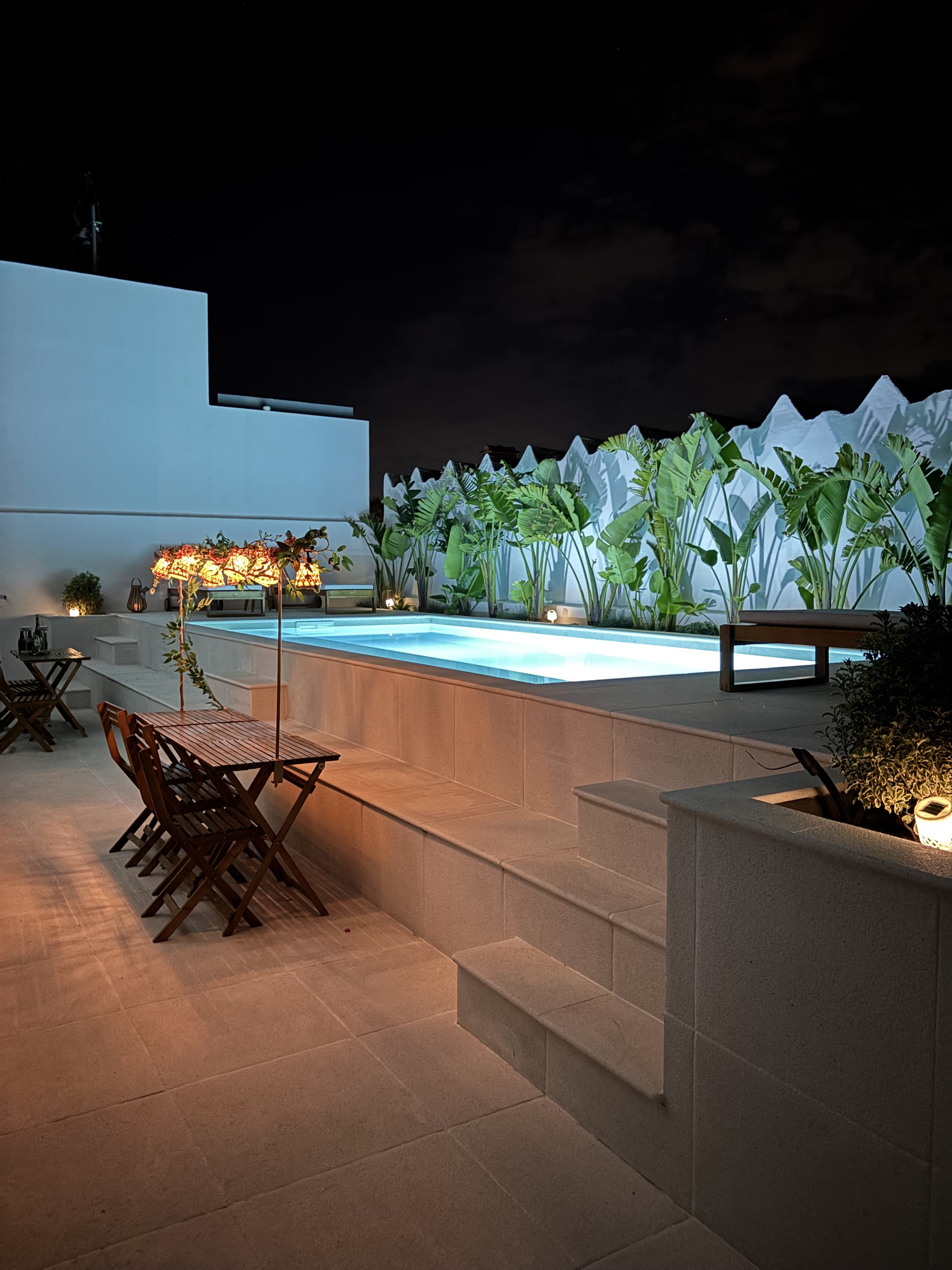 Night view of an outdoor illuminated swimming pool with surrounding seating, plants, and a black sky.