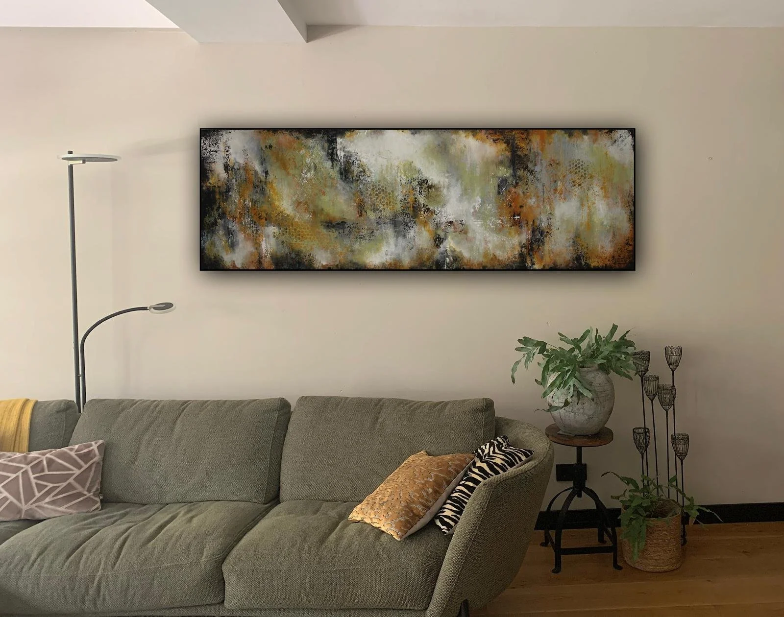 Woonkamer met beige bank, abstract kunstwerk aan de muur, groene planten, vloerlamp en sierkussens.