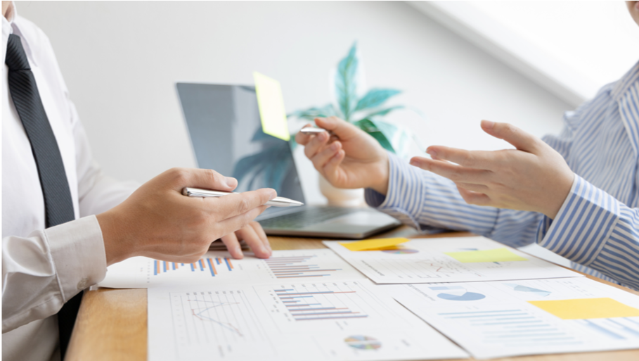 Two people having a business discussion at a desk with documents and charts, one person is writing on a notepad, the other is gesturing.