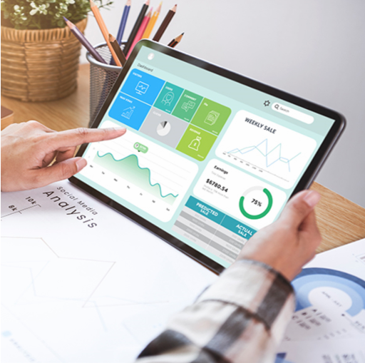 Person using a tablet displaying analytics and sales data with various charts, graphs, and icons, on a wooden desk with papers and a pen holder with colored pencils.