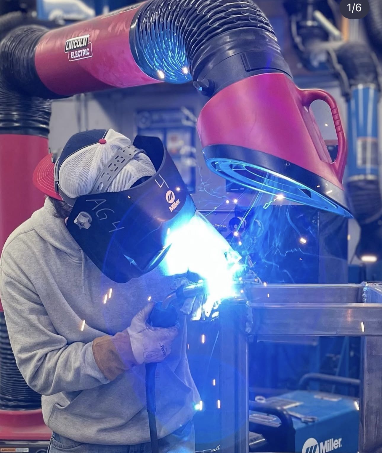 A person welding metal with a welding helmet, sparks and bright blue light, in a workshop with industrial equipment.
