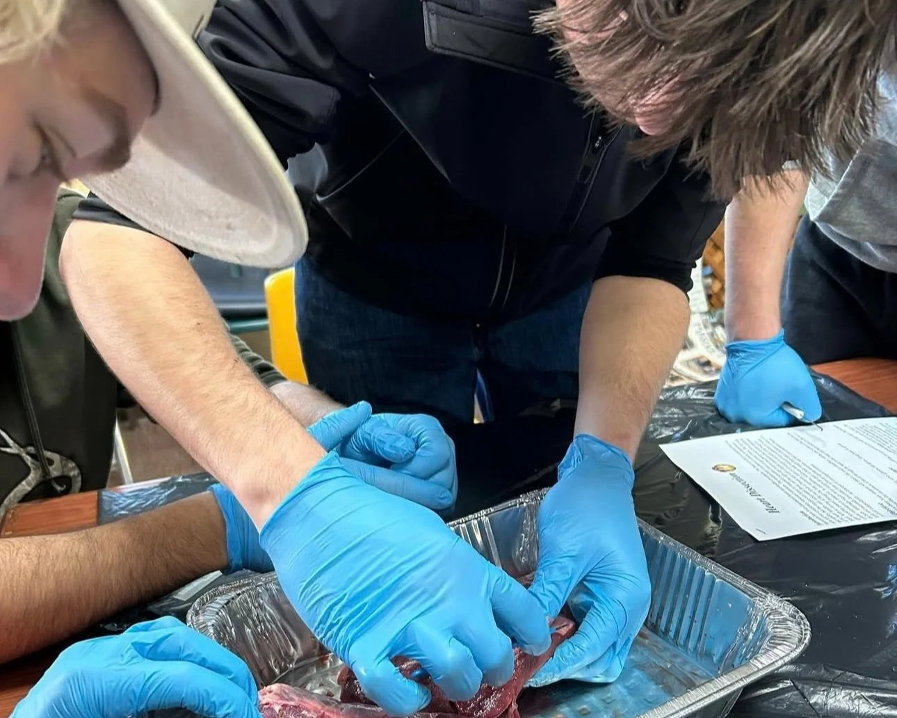 People wearing blue gloves handling and preparing meat in a metal tray, with one person wearing a white hat and others assisting, in a setting that appears to be a food handling or training environment.