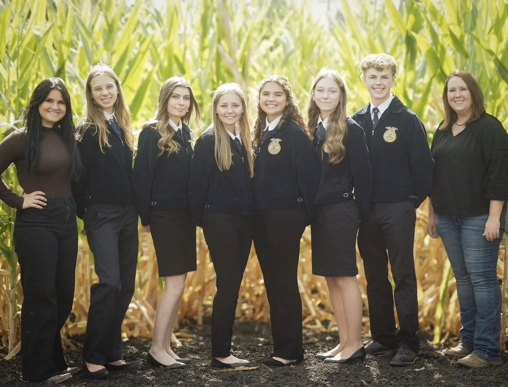 Group of seven young people standing outdoors in front of tall corn plants, dressed in casual and school clothing, smiling at the camera.