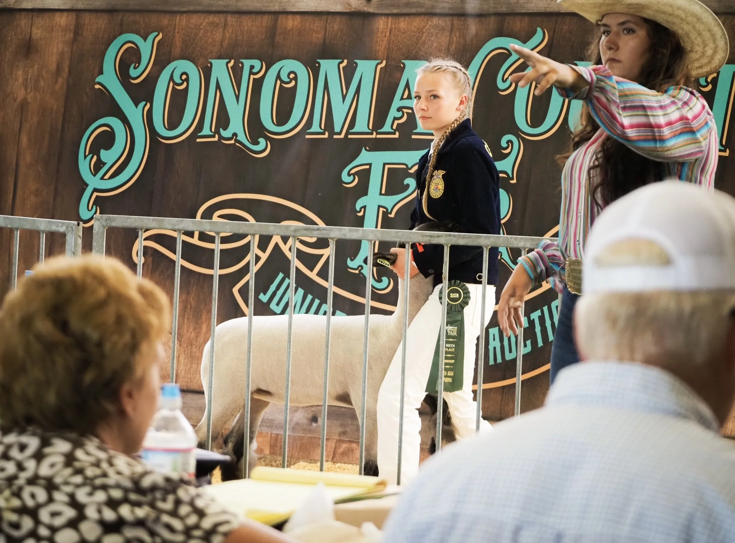 Two young girls standing behind a metal barrier at a fair or livestock show, with one girl wearing a cowboy hat and the other with braided hair, in front of a brown wooden wall with decorative teal lettering.