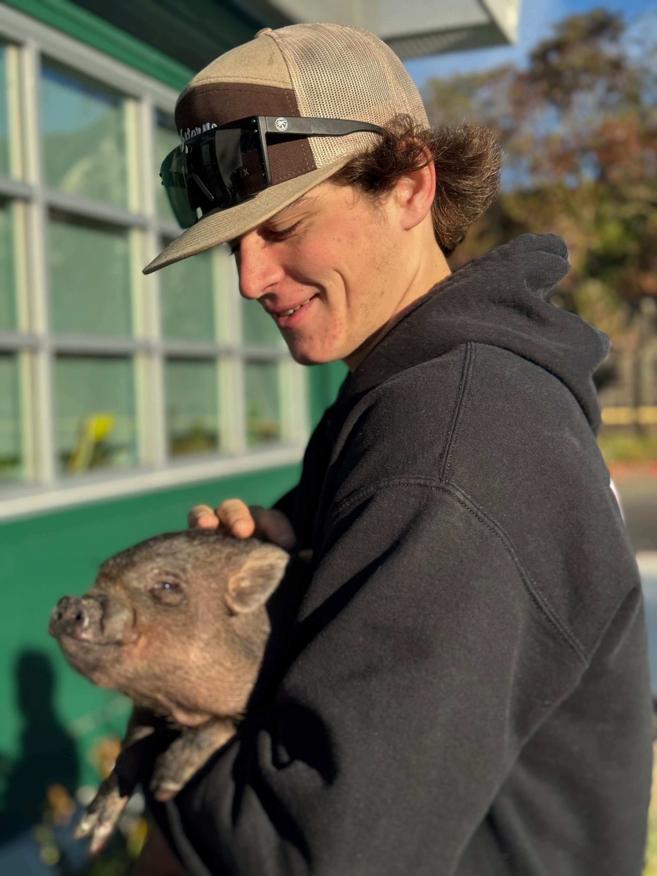 A young man wears a beige cap with a mesh back, sunglasses on the cap, and a black hoodie. He is smiling and holding a piglet in his arms outdoors.