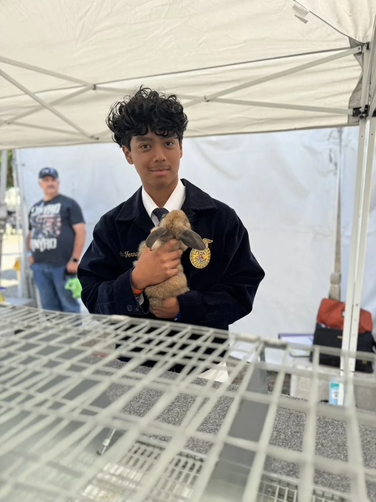 A young man in a black jacket holding a small rabbit in front of a white tent at an outdoor event, with another person standing in the background.