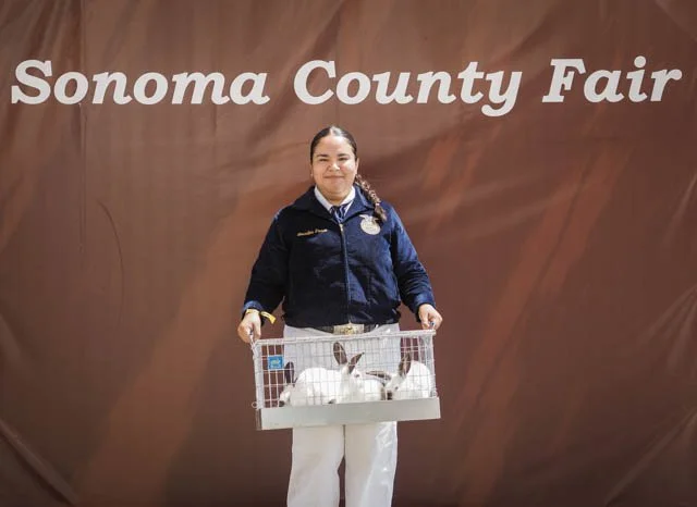 Young girl smiling while holding a bunny cage at the Sonoma County Fair with a brown backdrop.