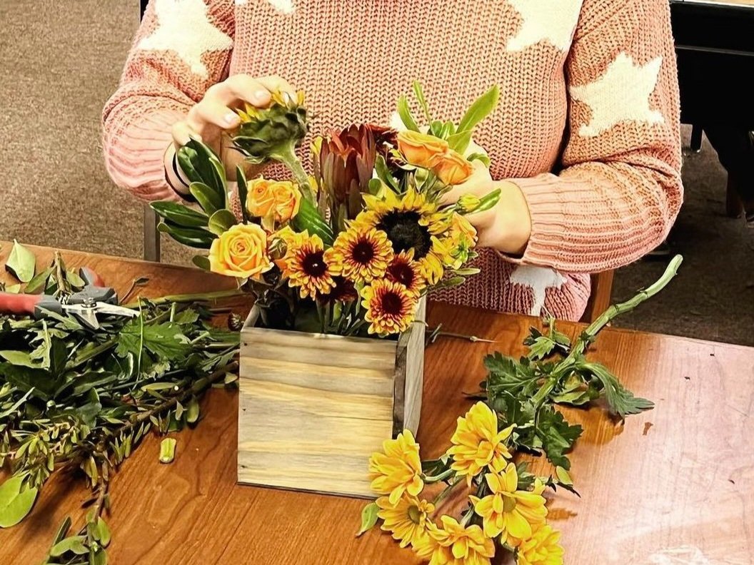 Person arranging a yellow and orange flower bouquet in a wooden box on a wooden table.
