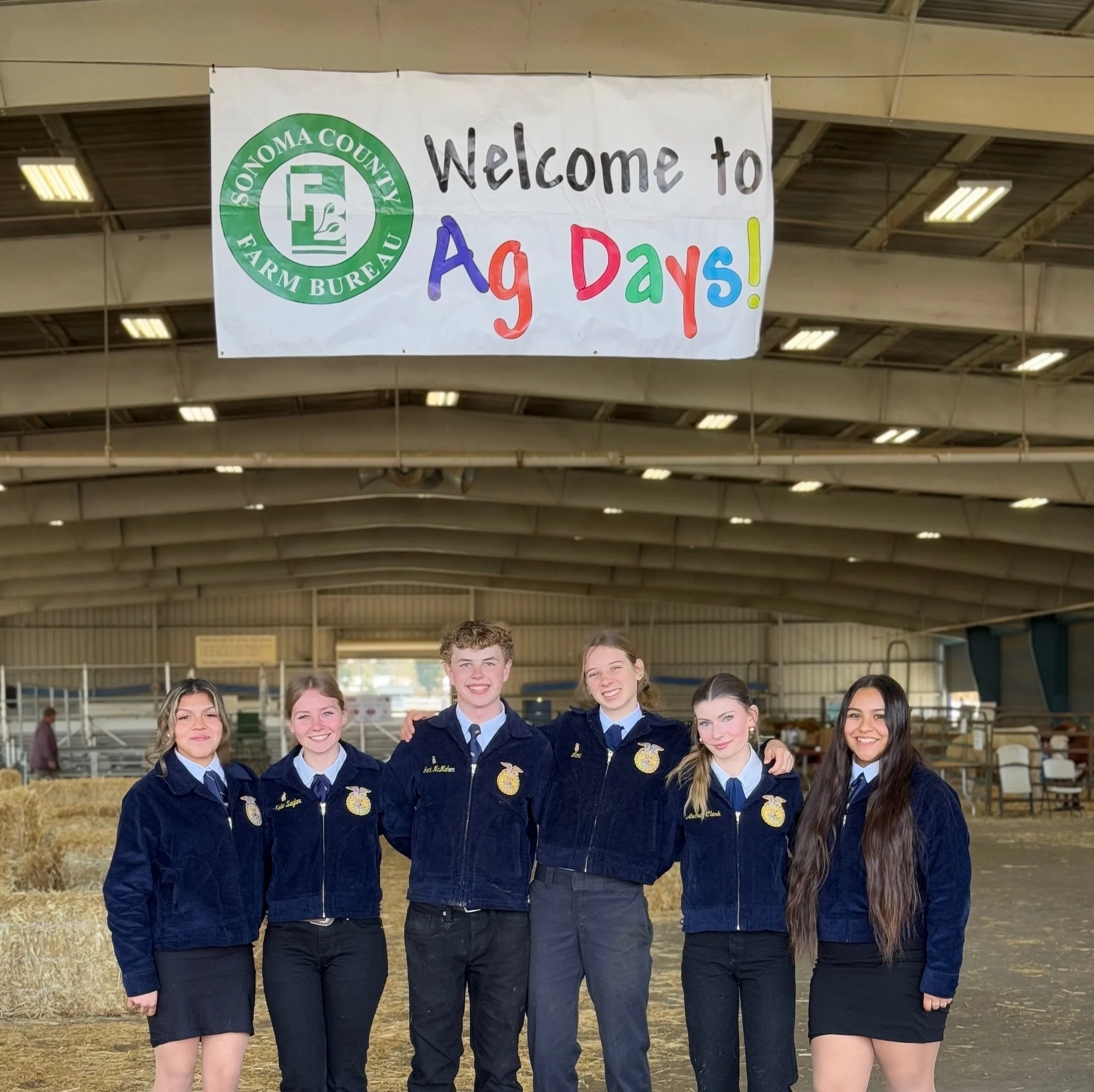 Group of young women and a young man in FFA jackets standing in a barn at an agricultural event.