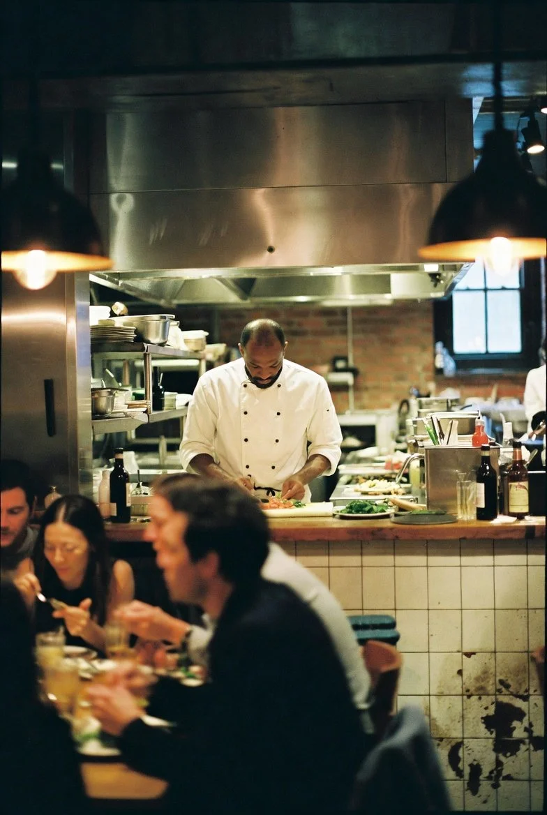 Chef preparing food in a busy restaurant kitchen with diners dining in the foreground.