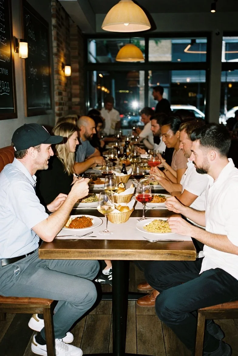 Group of people dining together in a restaurant, sitting at a long table with plates of food, glasses of wine, and baskets of bread, with a warm indoor ambiance and large windows in the background.