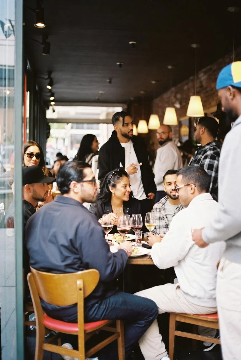 A group of diverse people enjoying a meal at a restaurant with drinks, seated around a table, while a waiter takes their orders. The restaurant has warm lighting and a cozy atmosphere.