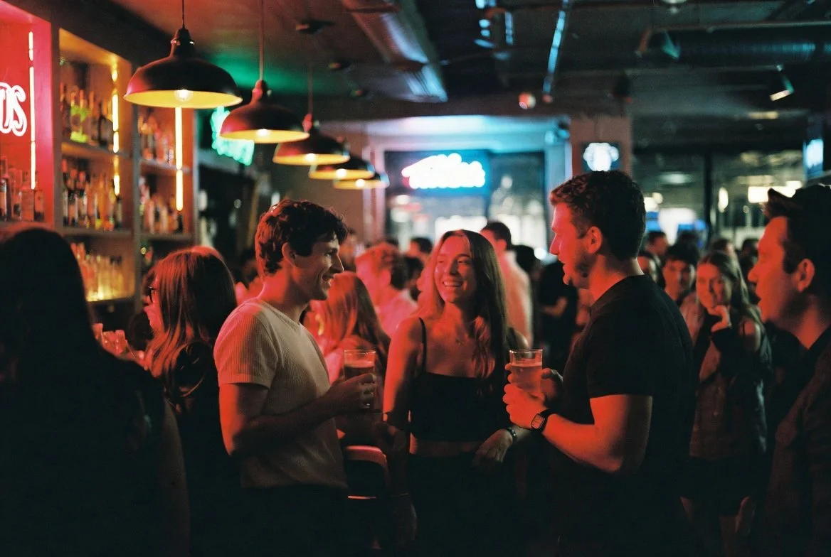 Group of young adults enjoying drinks and socializing in a bar with dim, colorful lighting.