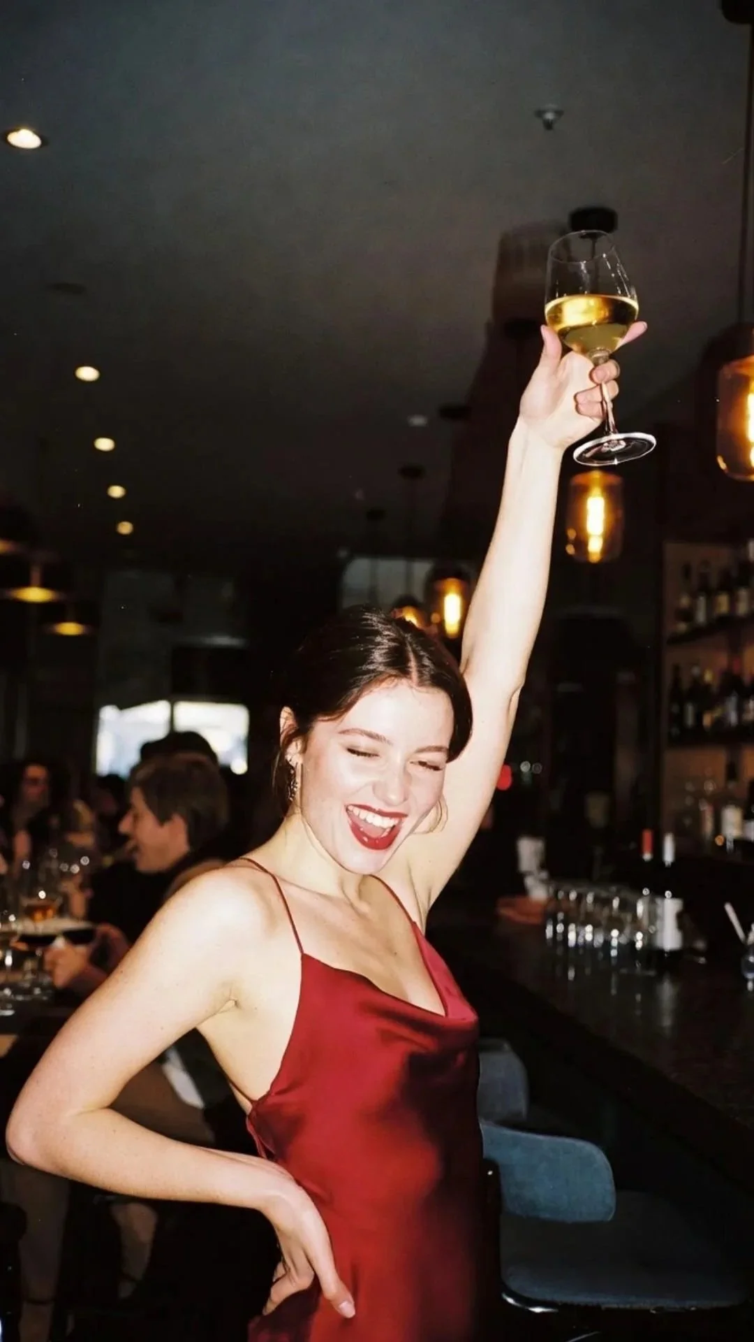 A woman in a red satin dress raising a glass of white wine and smiling in a bar or restaurant setting.