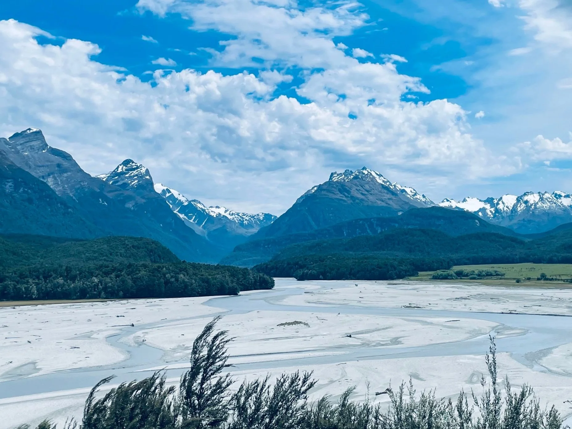Snow-covered river valley surrounded by green forests and tall mountains with snow-capped peaks under a partly cloudy blue sky.