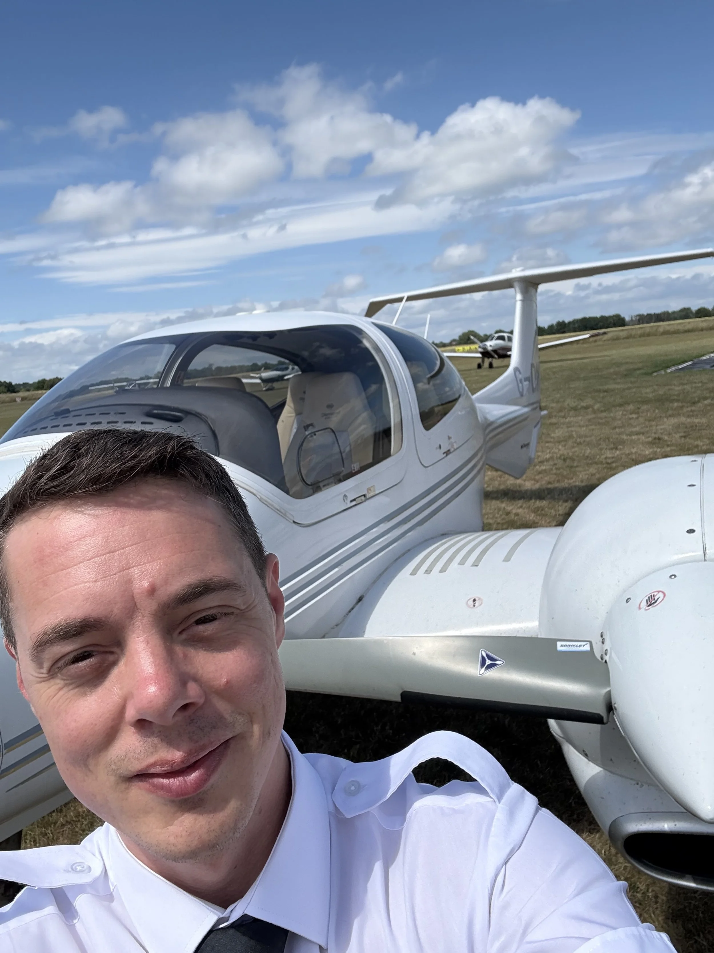 A smiling man in a white shirt taking a selfie in front of a small white aircraft on a grassy airfield under a partly cloudy sky.