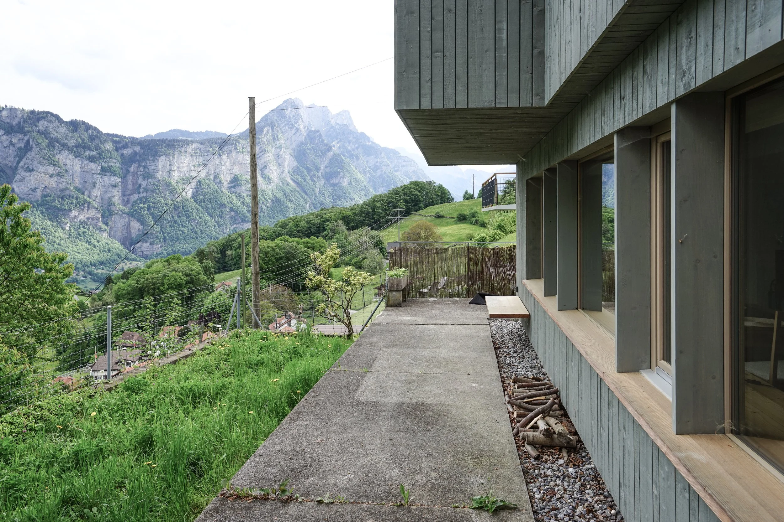 Blick von einer Terrasse an einem modernen Haus mit Holzfassade auf grüne Hügel und Berge im Hintergrund.
