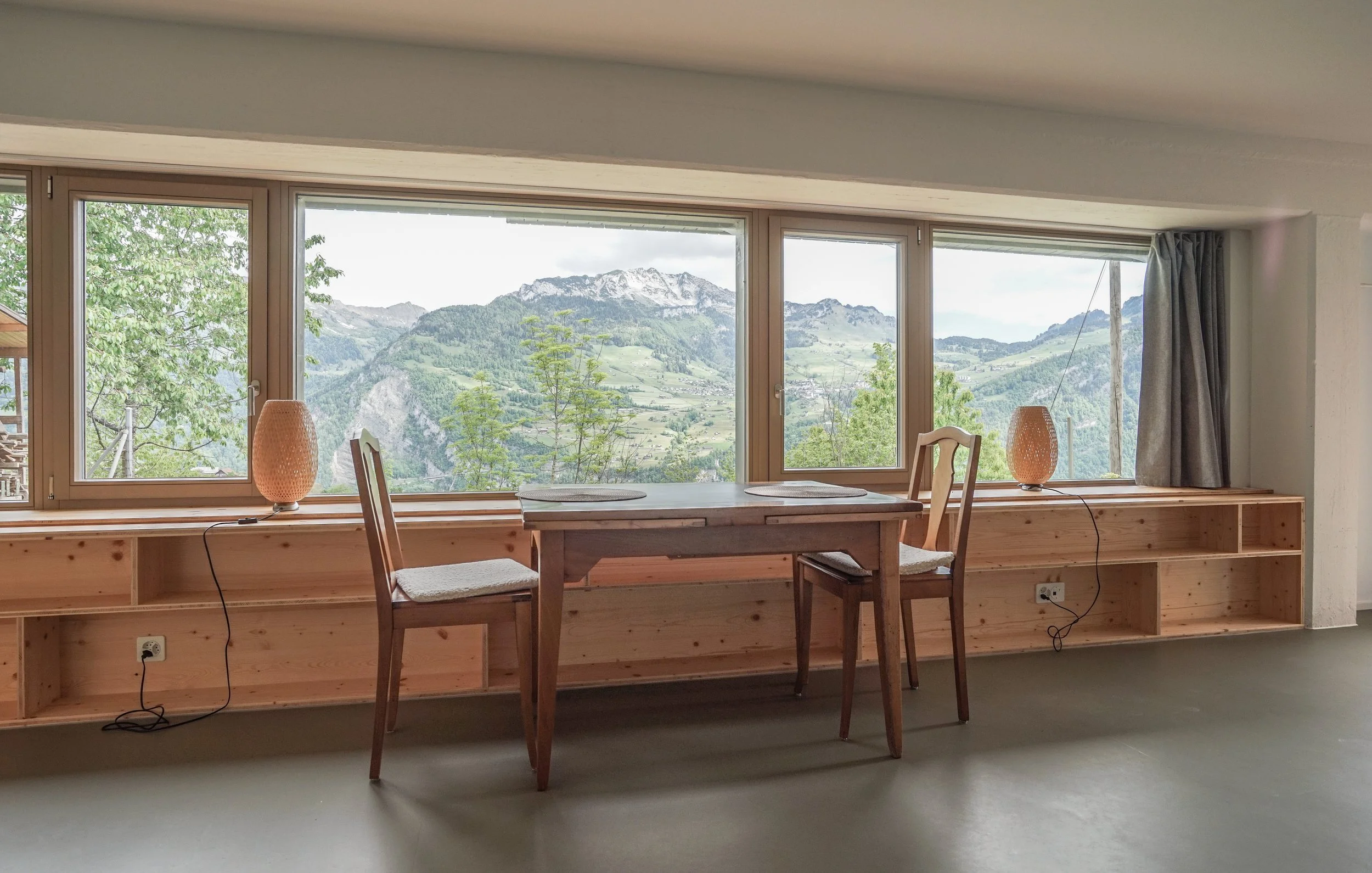 Blick durch große Fenster auf eine Berglandschaft mit grünen Hügeln und schneebedeckten Gipfeln. Innenraum mit Holzmöbeln und zwei Lampen, eine auf der Fensterbank, die andere auf einem Stuhl, neben einer hölzernen Tisch mit zwei Stuhlen.