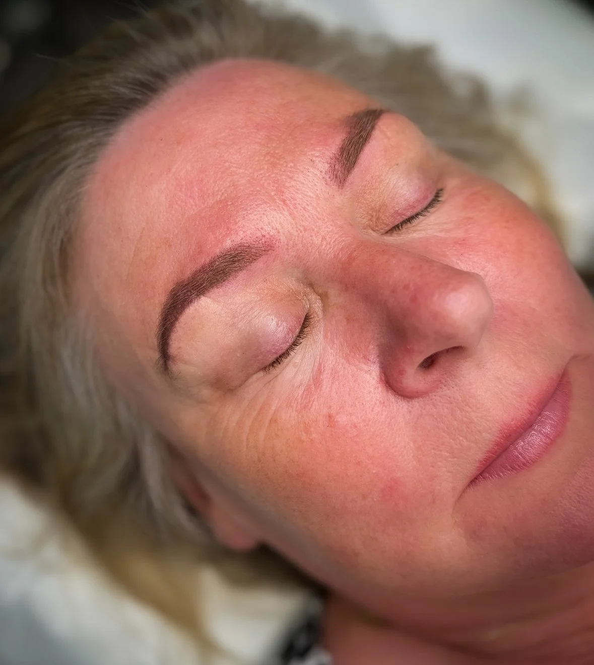 Close-up of a woman with freshly tattooed or microbladed eyebrows, lying down with her eyes closed.