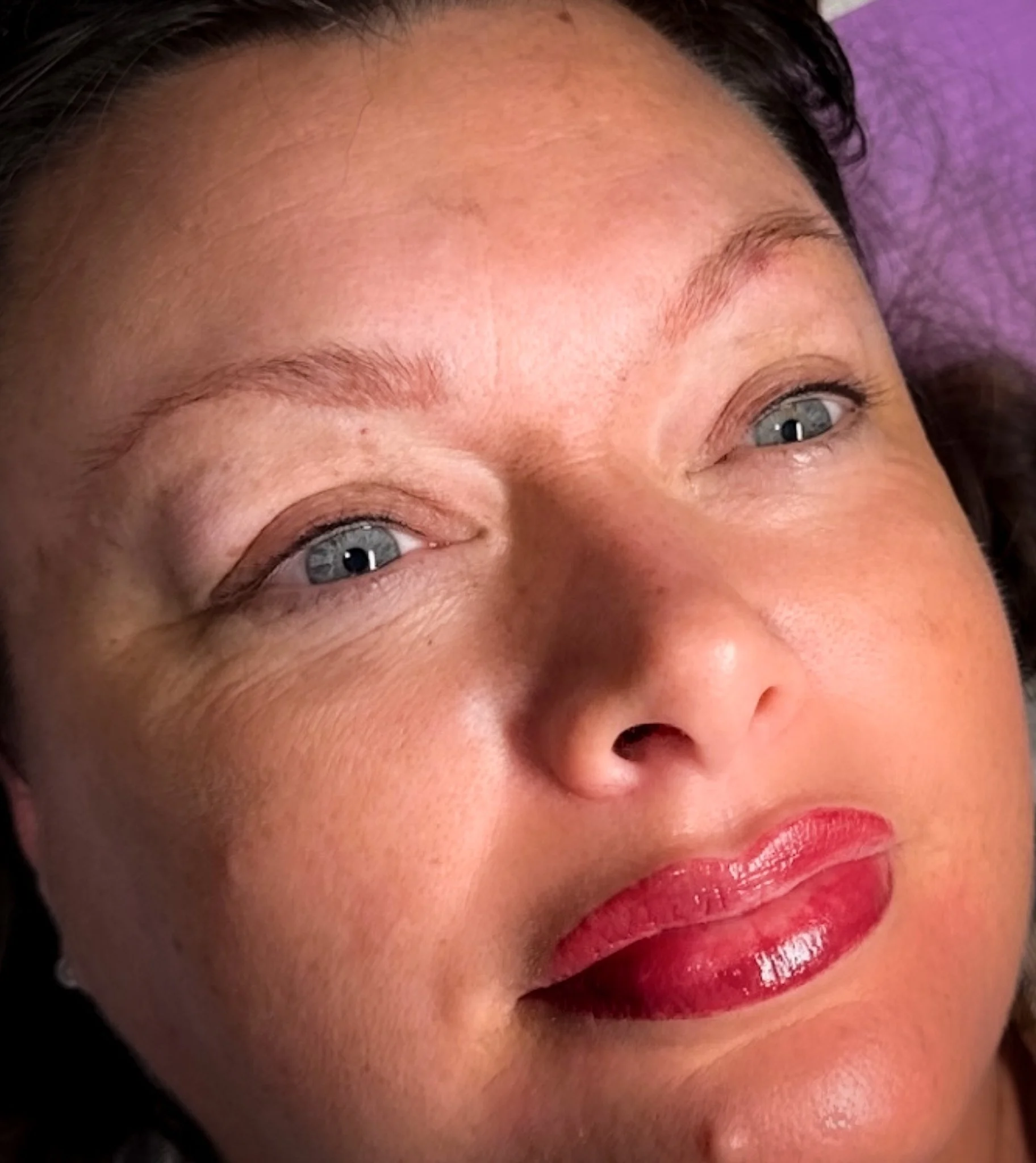 Close-up photo of a woman with blue eyes, light skin, red lipstick, and brown hair lying down on a purple surface.