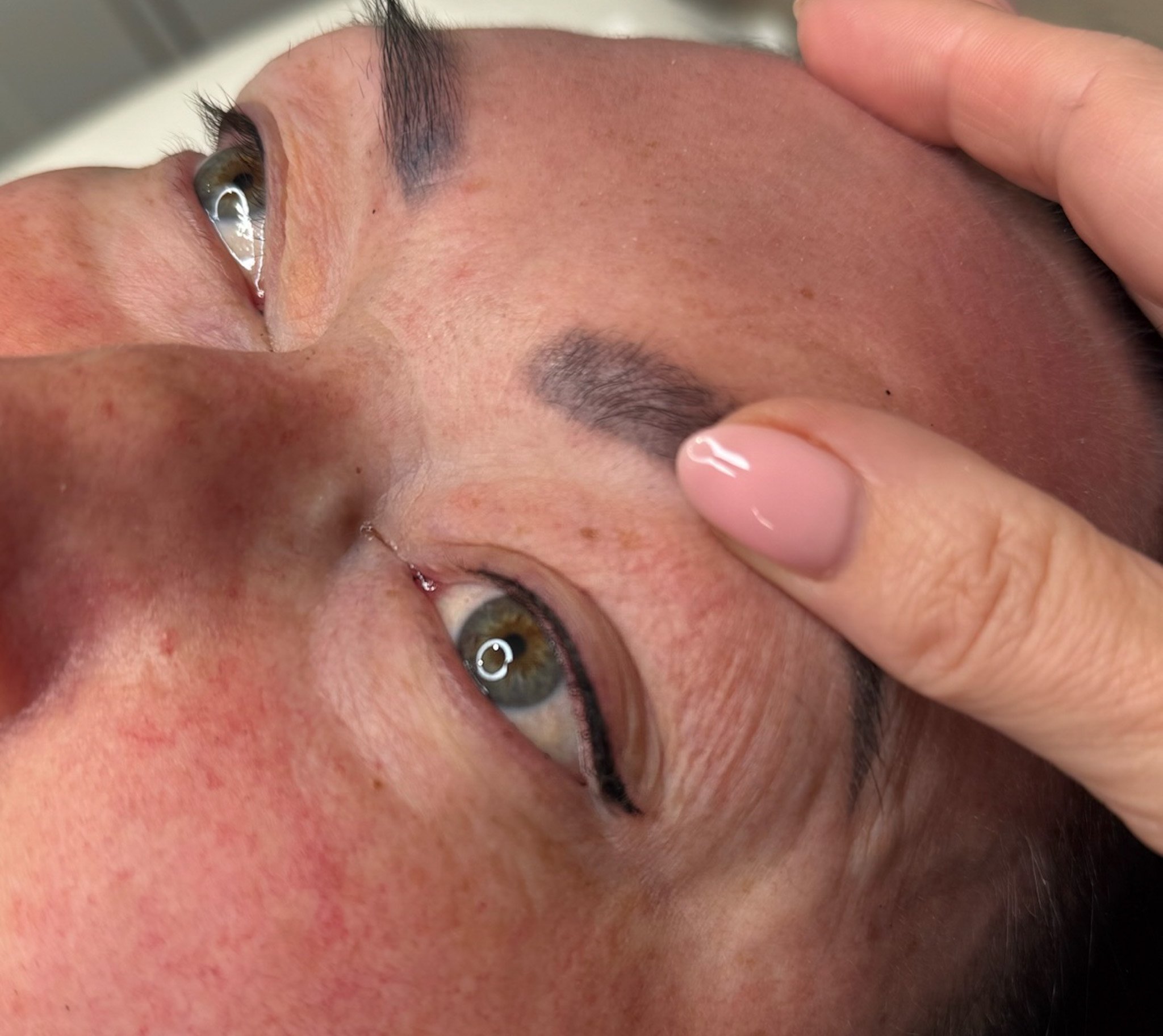 A close-up of a person receiving eyebrow microblading or cosmetic tattooing, with a technician holding the person's forehead.