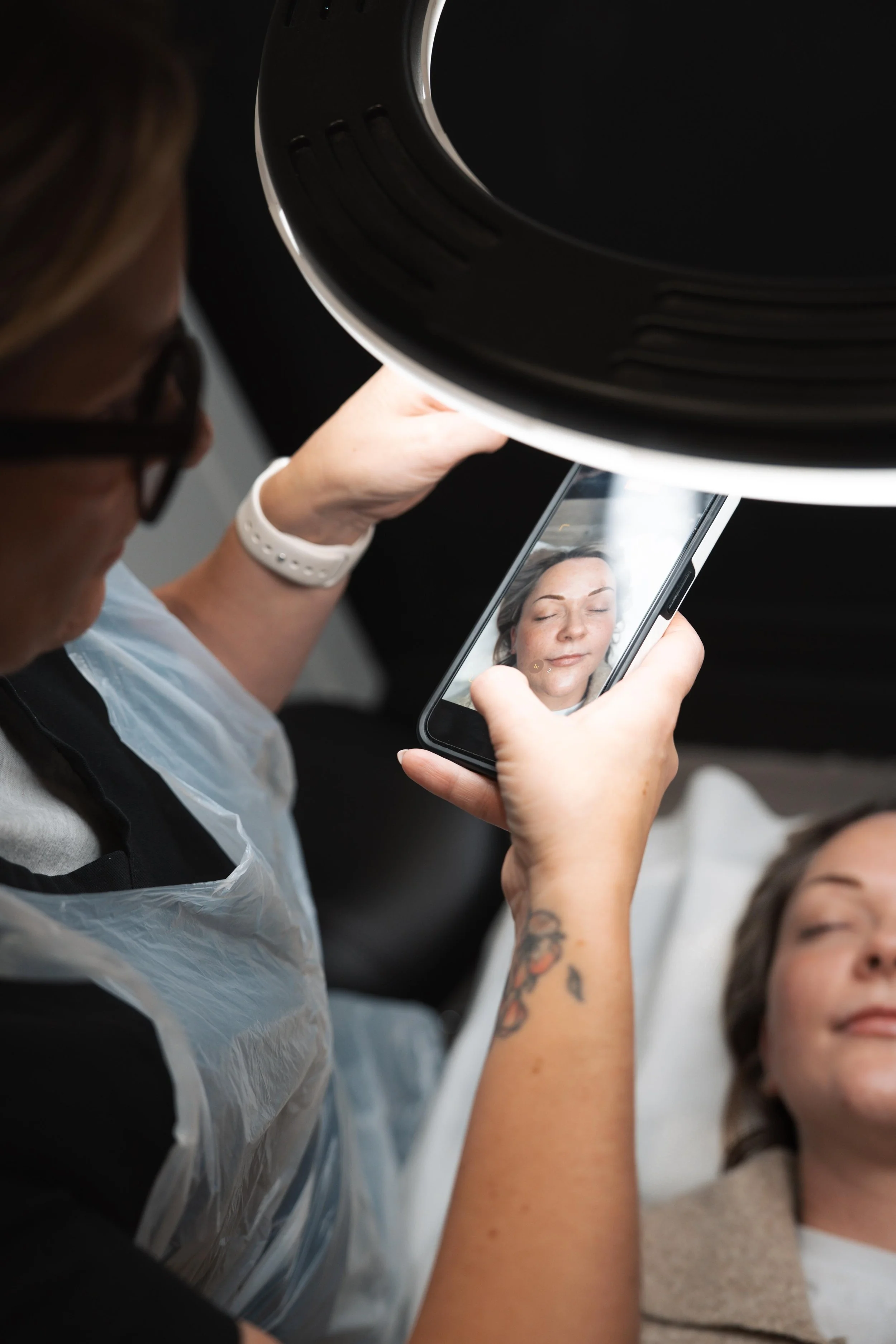 A woman taking a selfie of her with a calm face in a medical or spa setting, with another woman lying down in the background.