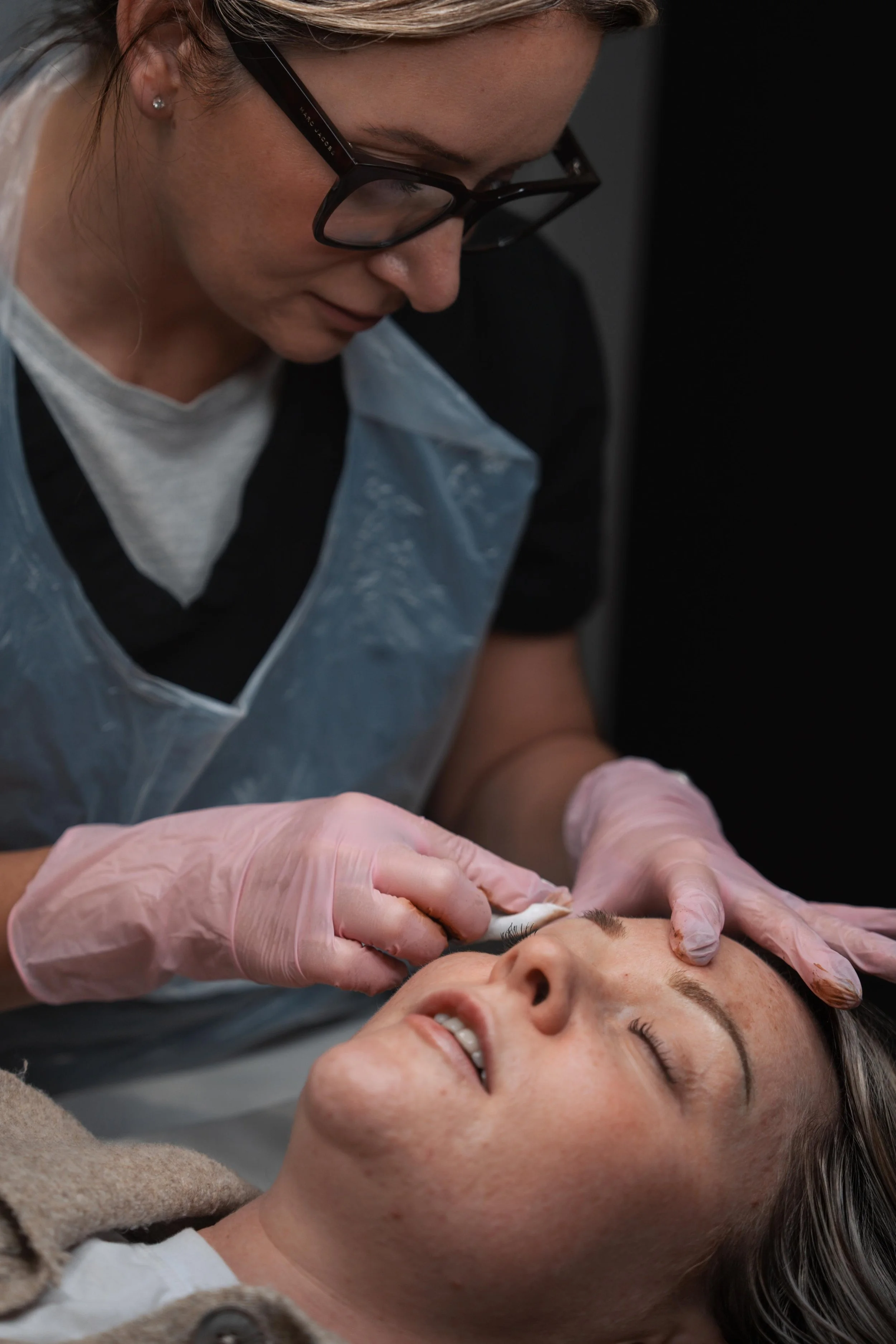 A woman receiving an eyelash extension procedure from a technician wearing gloves and protective glasses in a clinical setting.