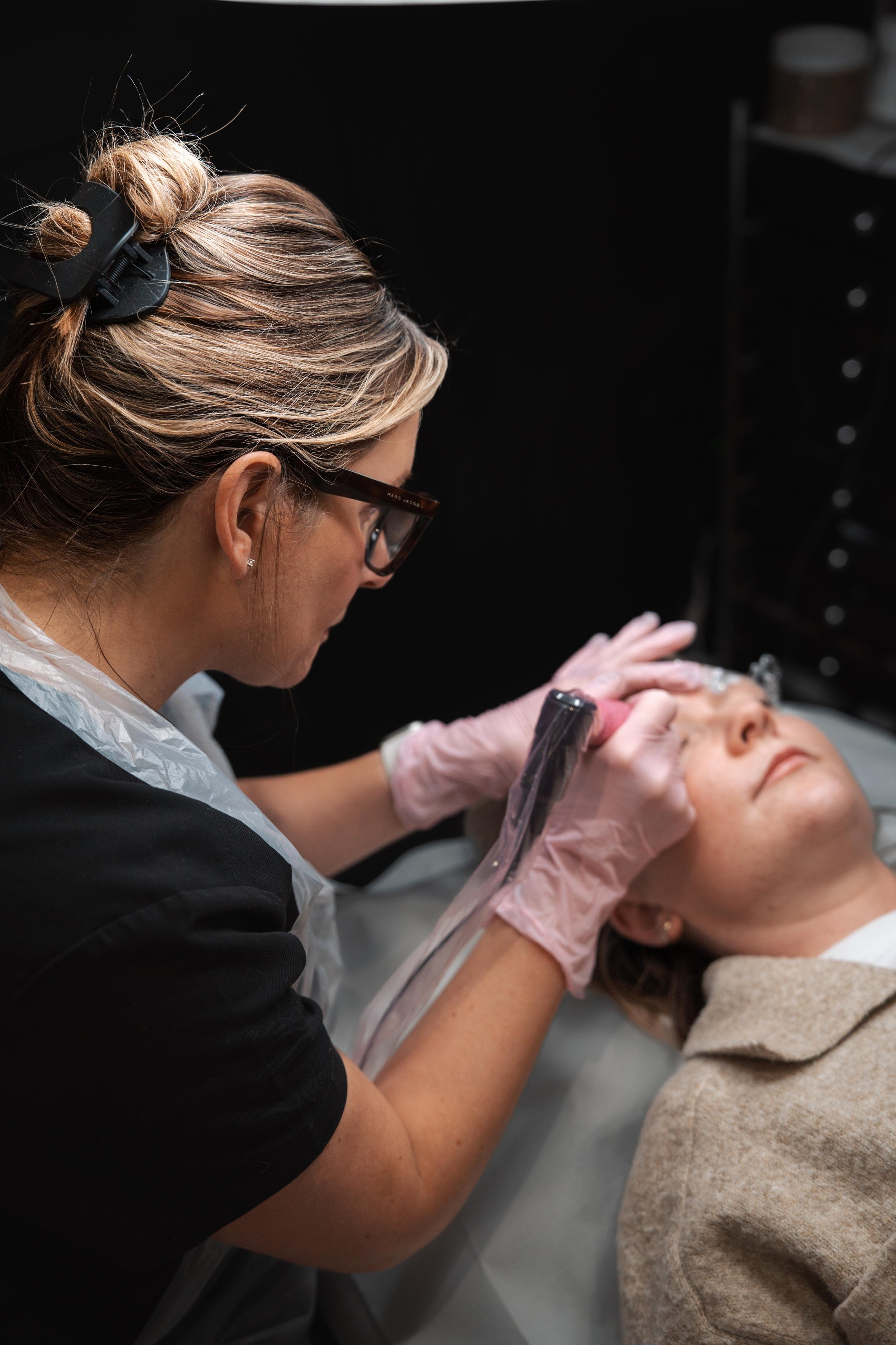 Skincare professional performing a facial treatment on a woman lying on a treatment bed, in a clinical setting.