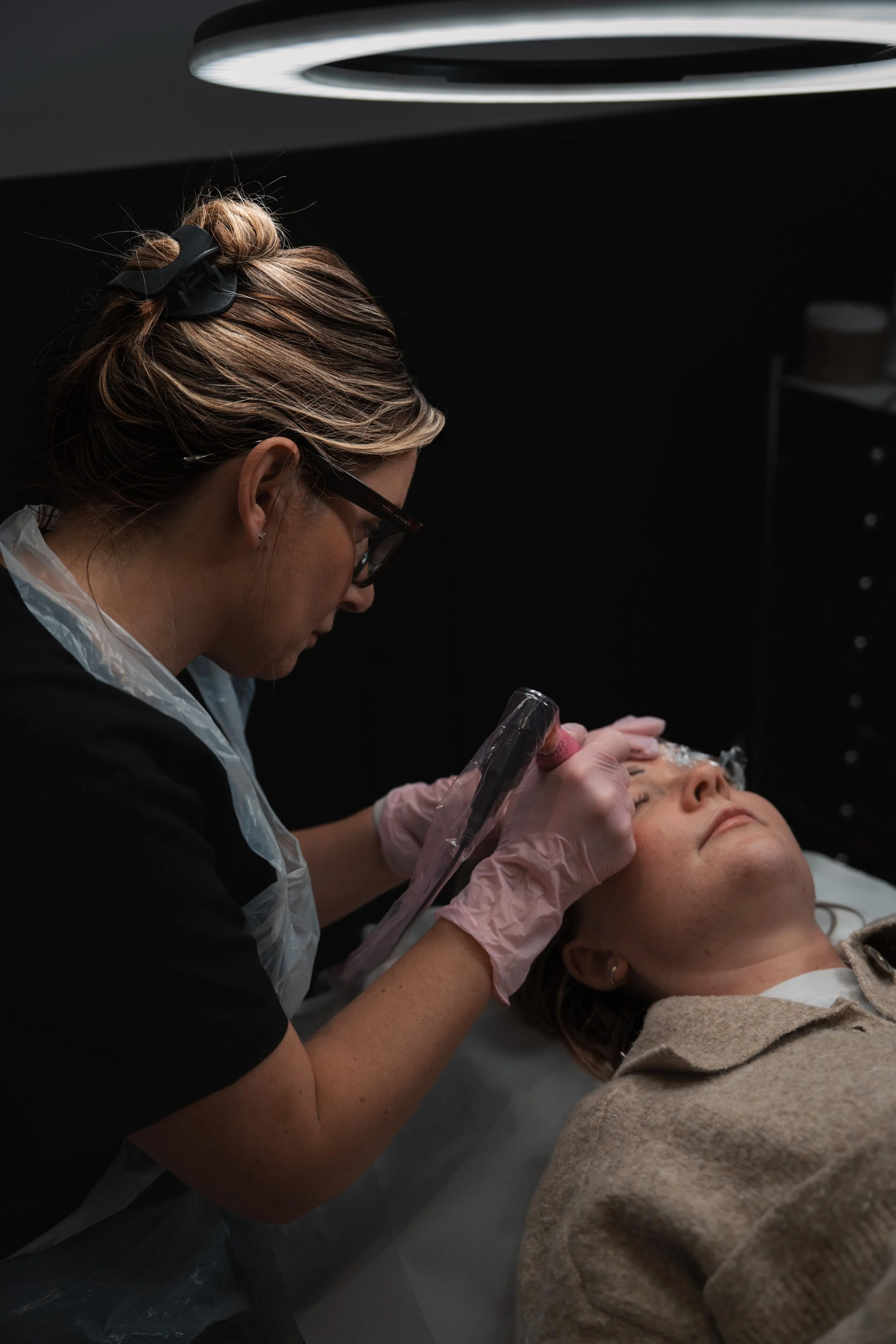 A woman performing a cosmetic or skincare procedure on another woman lying down with her eyes closed inside a clinic or treatment room.
