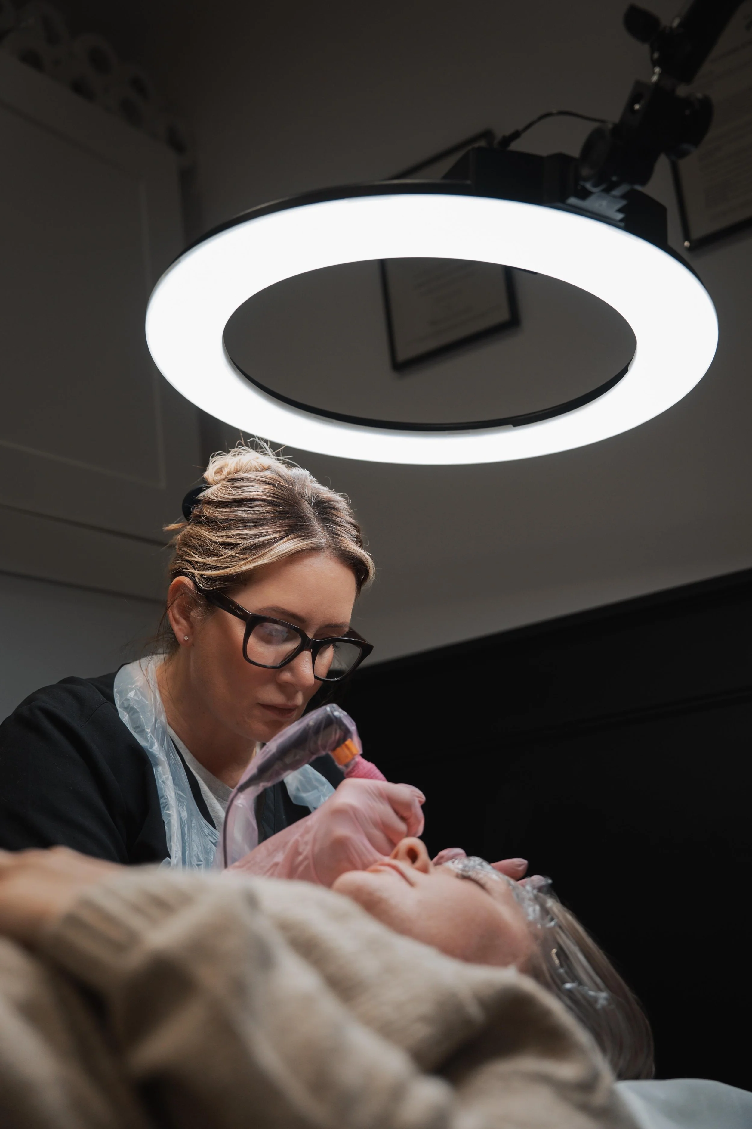 A medical professional in black scrubs and glasses performs a procedure on a patient lying down, under bright lighting.