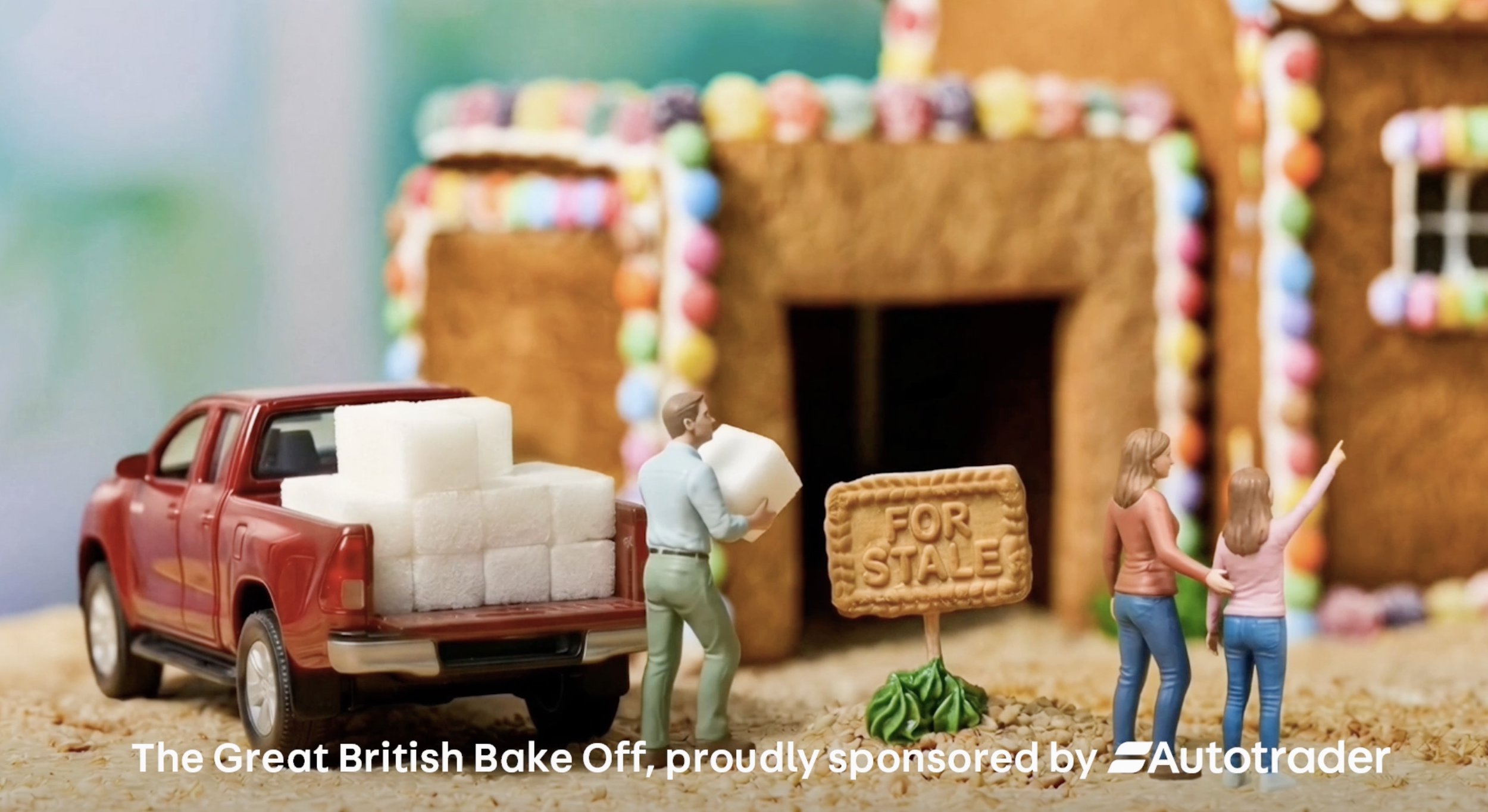 Miniature scene with toy figures of a man, two women, and a red toy truck carrying marshmallow treats in front of gingerbread houses decorated with colorful candies.