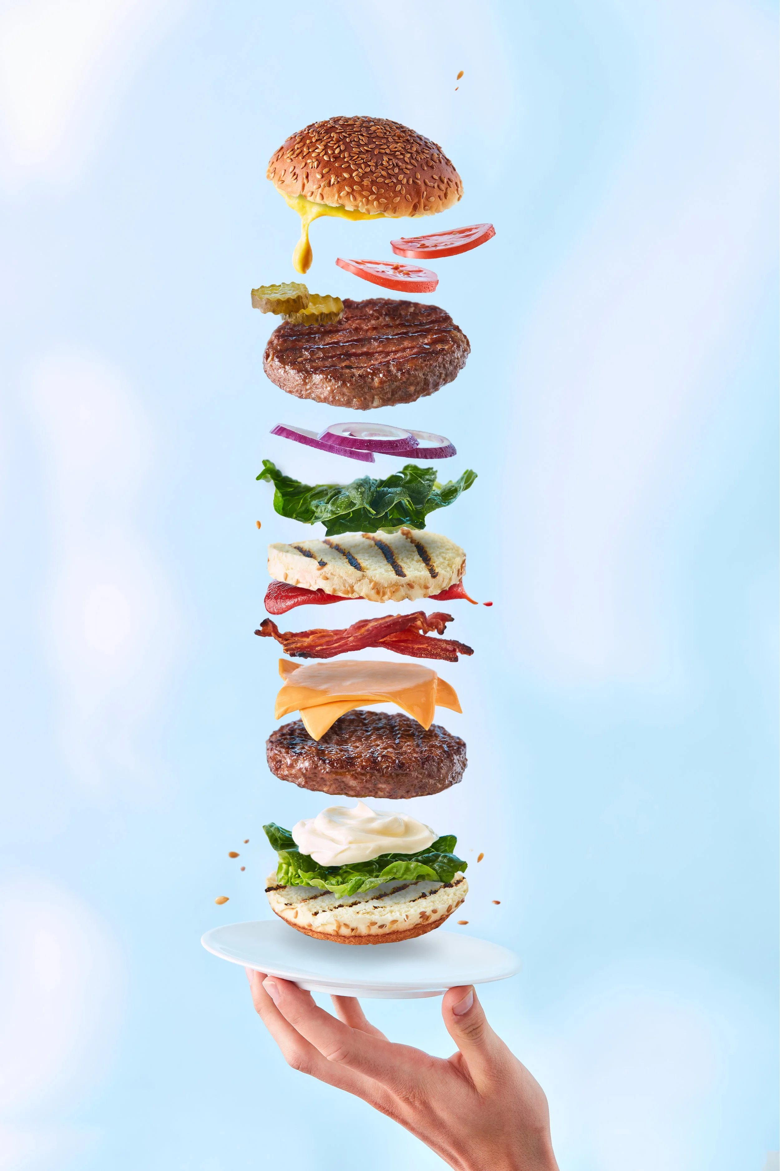 A hand holding a white plate with a stack of various burger ingredients suspended in mid-air against a light blue background, showing lettuce, grilled burger patties, sliced tomatoes, onions, pickles, cheese, and sesame seed bun.