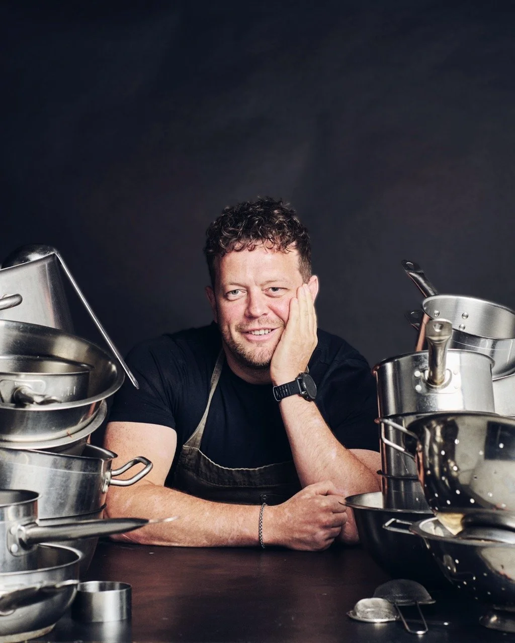 A smiling male chef with curly hair, wearing a black shirt and apron, inside a kitchen with stainless steel pots and pans.