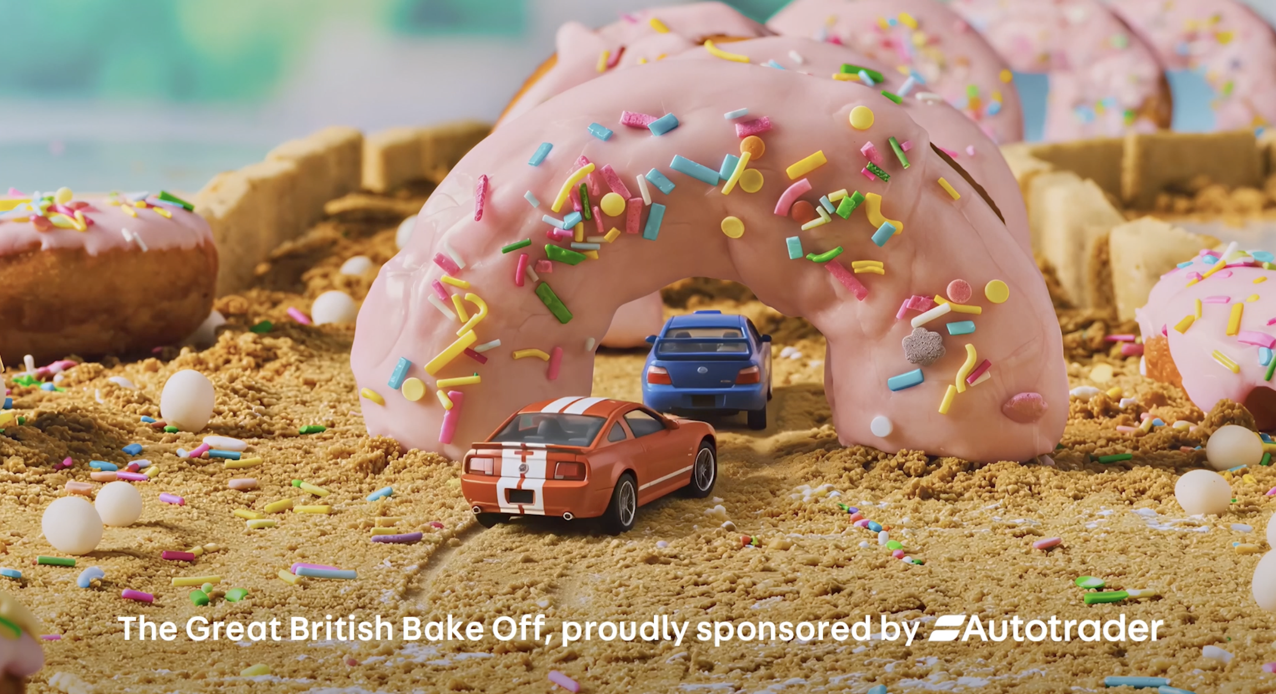 A creative scene with a large pink frosted donut decorated with colorful sprinkles, surrounded by smaller donuts, toy cars, and white balls on a sandy surface, with blurred background.