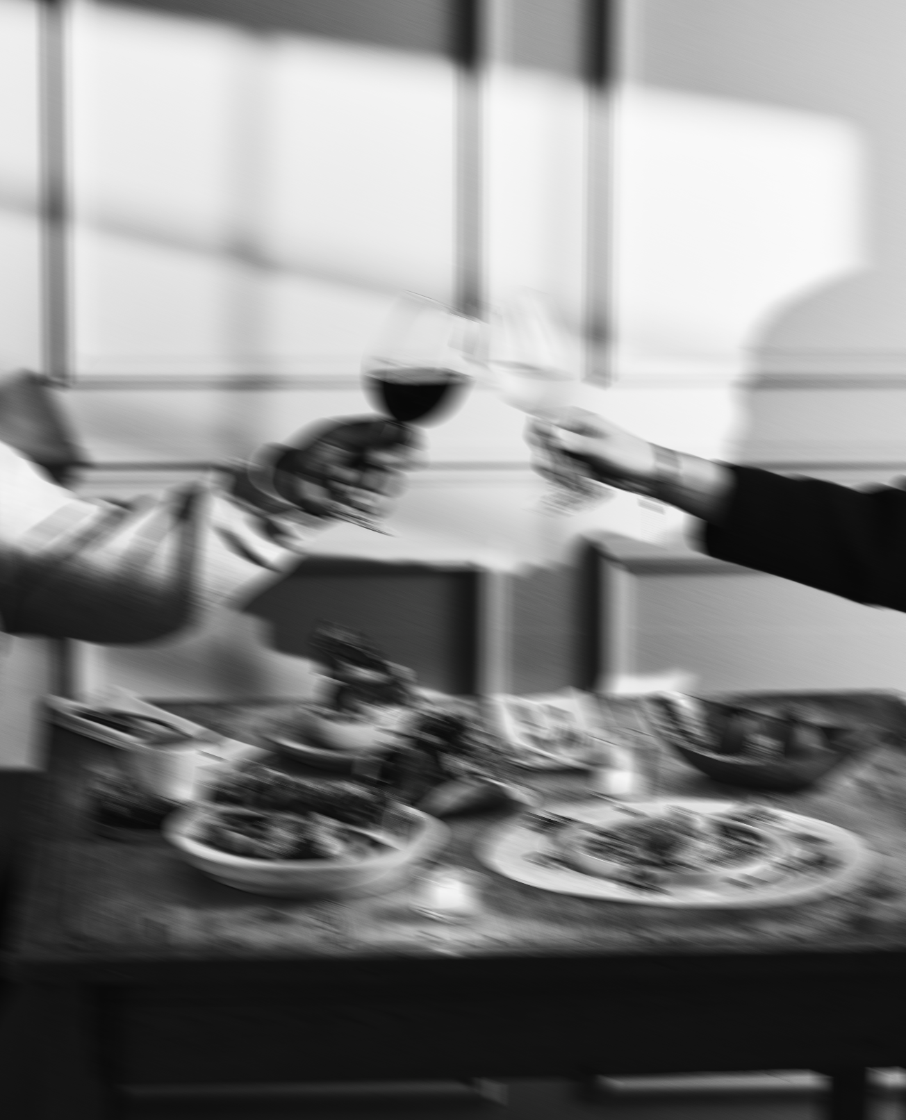 Blurred black and white photo of a wine glass in a person's hand, with a rack of dishes below and shelves in the background.