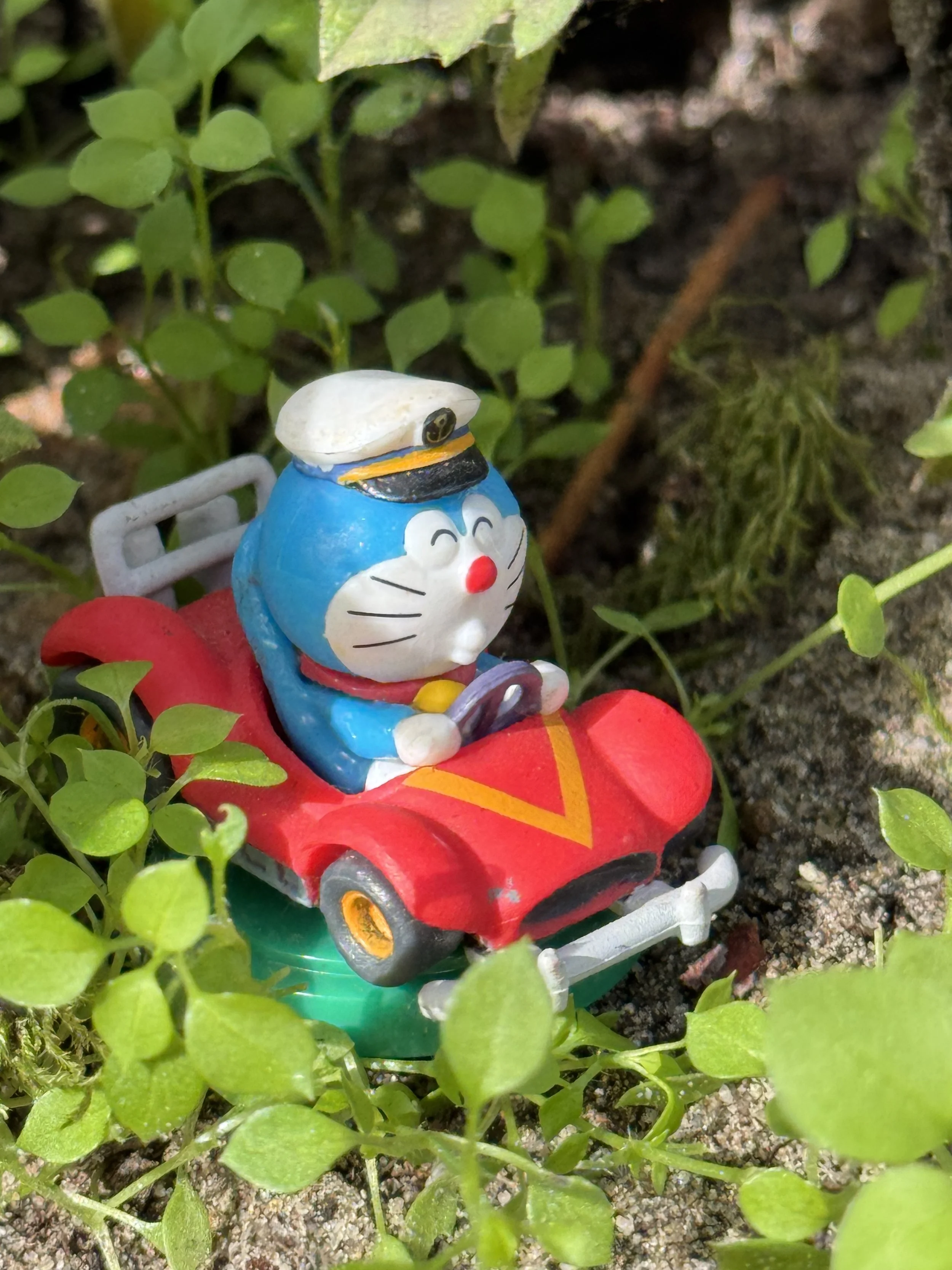 A small toy figure of Doraemon, a blue robotic cat from Japanese manga, is riding a tiny red go-kart in a patch of green plants and soil.