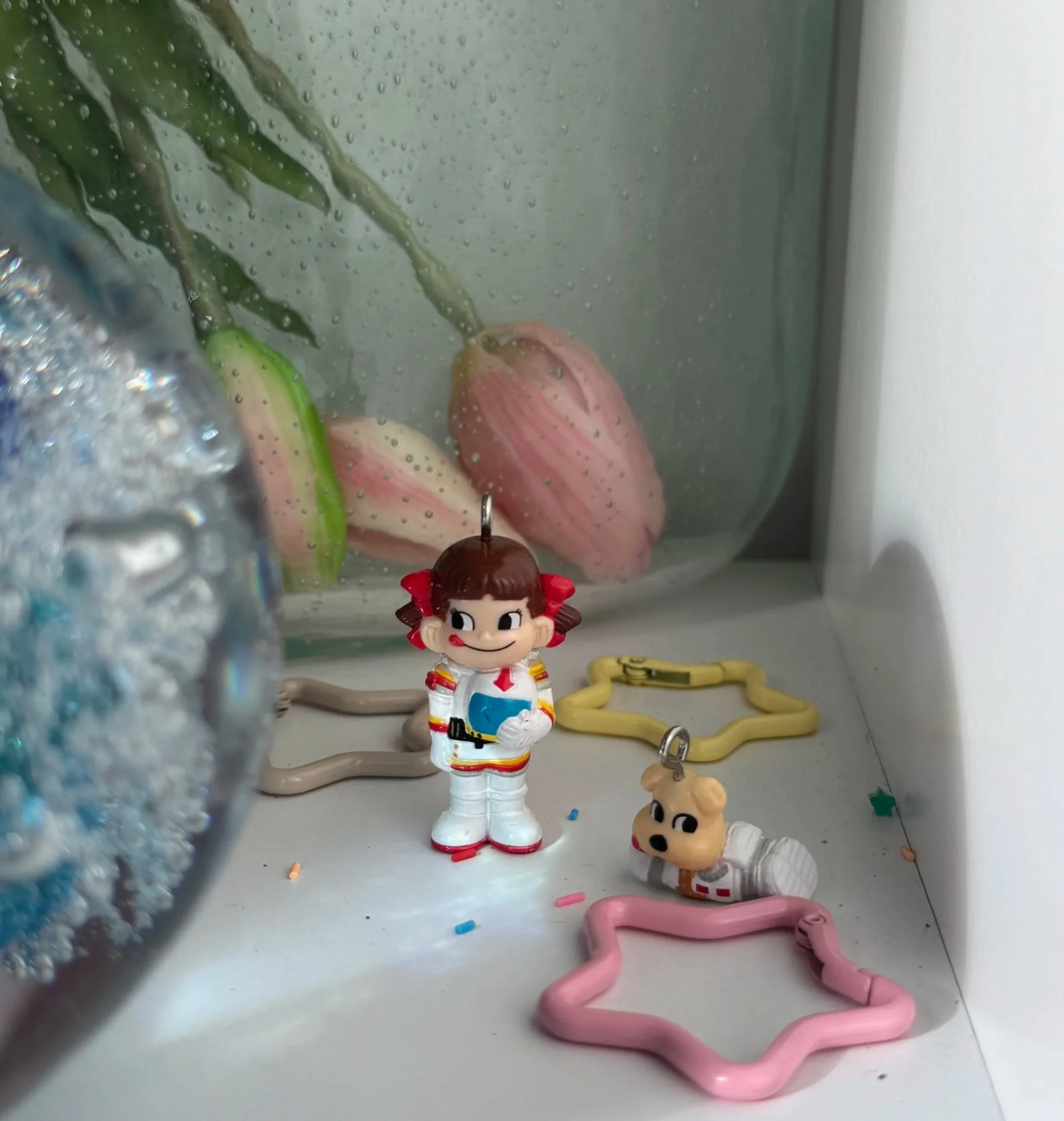 Small figurines of cartoon characters, including a girl with pigtails, a dog in a space suit, and plastic star-shaped molds, are placed on a white surface with a foggy glass container filled with pink tulips and green leaves in the background.