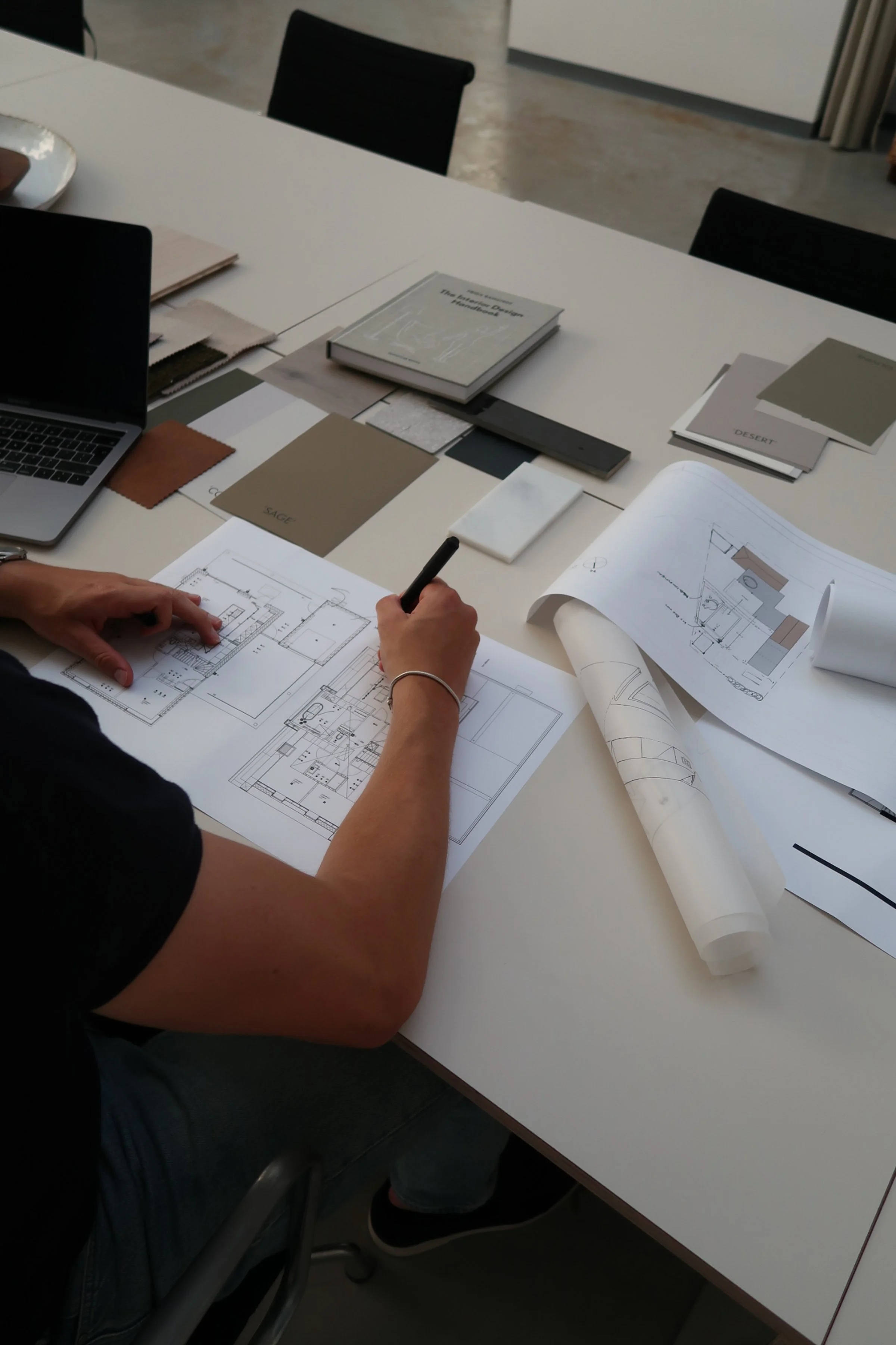 Een persoon bestudeert bouwtekeningen en interieurontwerpen op een tafel, omringd door kleur- en materiaalkleuren en boeken over ontwerp.