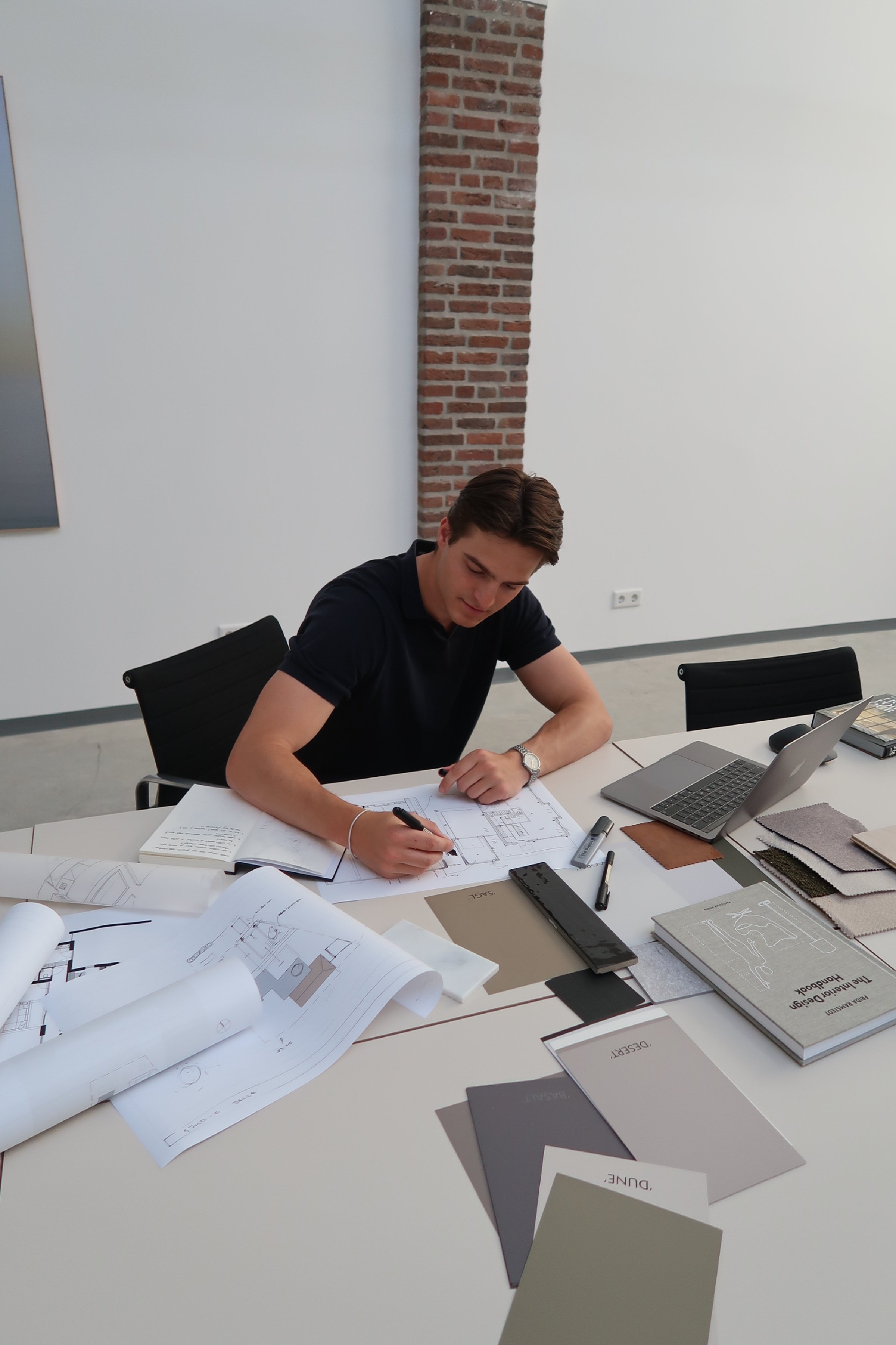 Man zittend aan een tafel met architectuurplannen, kleurstalen, een laptop en tekengerei in een vergaderruimte.