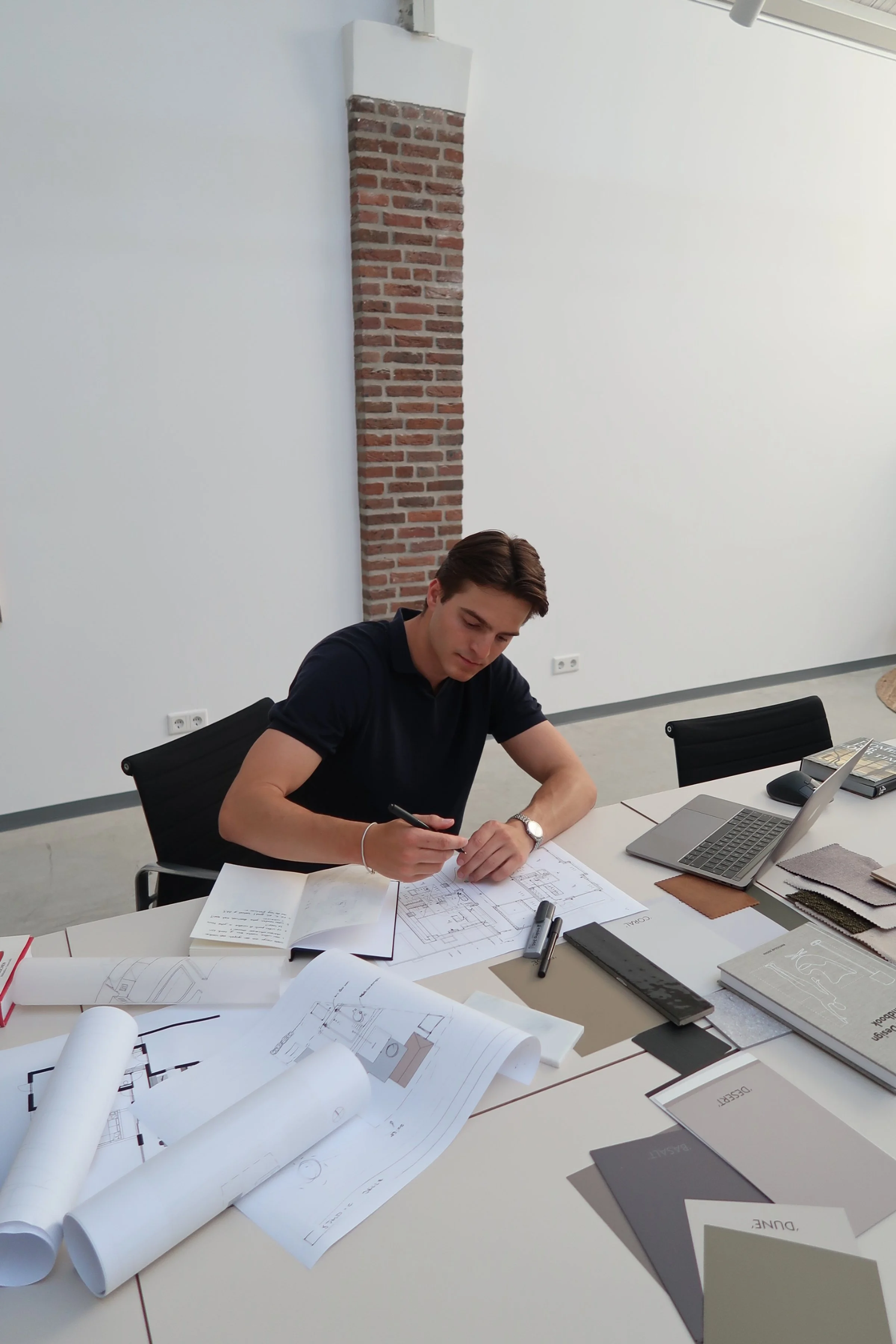 Man werkt aan bouwplannen op een bureau met blauwdrukken, een laptop, notitieboekjes, en stiften in een moderne kantoorruimte.