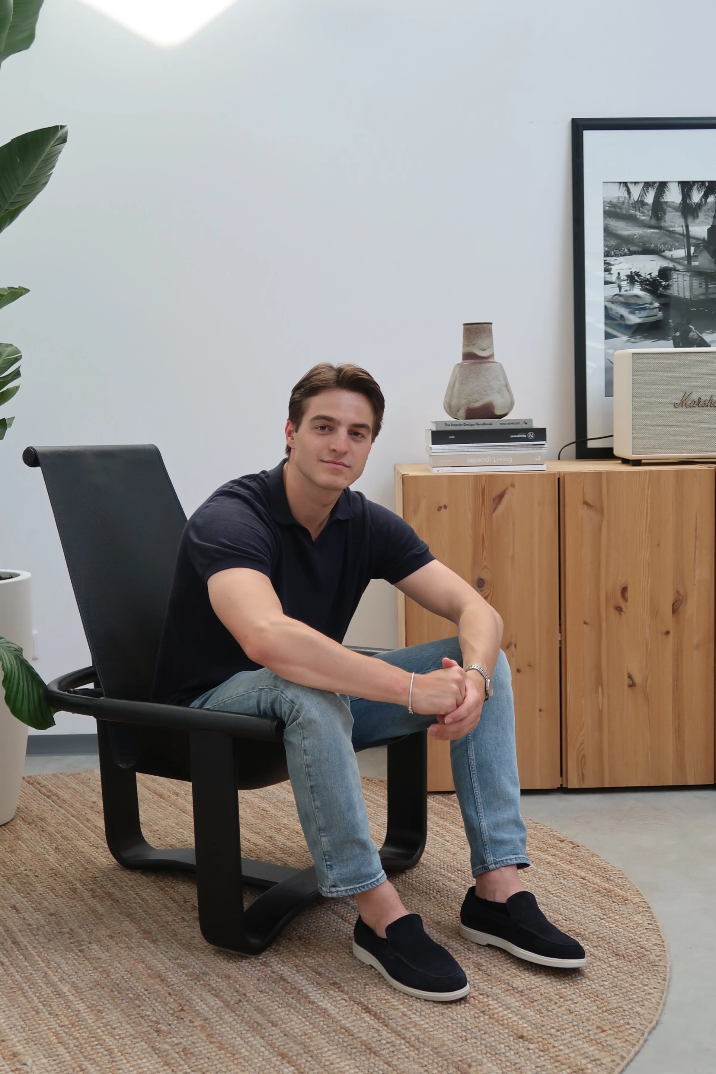 Jonge man zittend op een zwarte stoel in een modern ingericht kantoor of woonkamer met houten kasten, een grote foto op de muur, en planten.